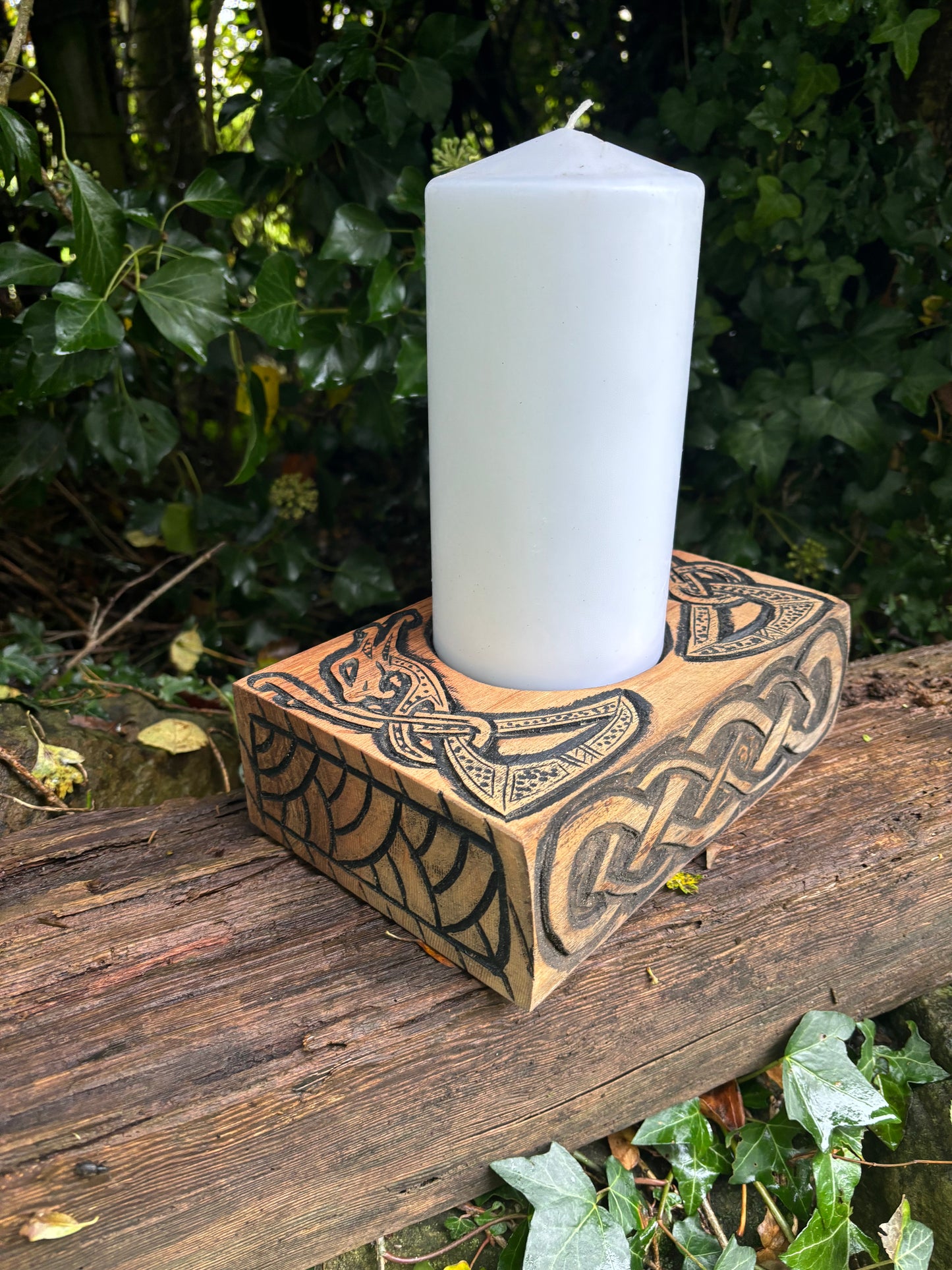White candle on carved wooden holder with intricate patterns, set on a log with green foliage.
