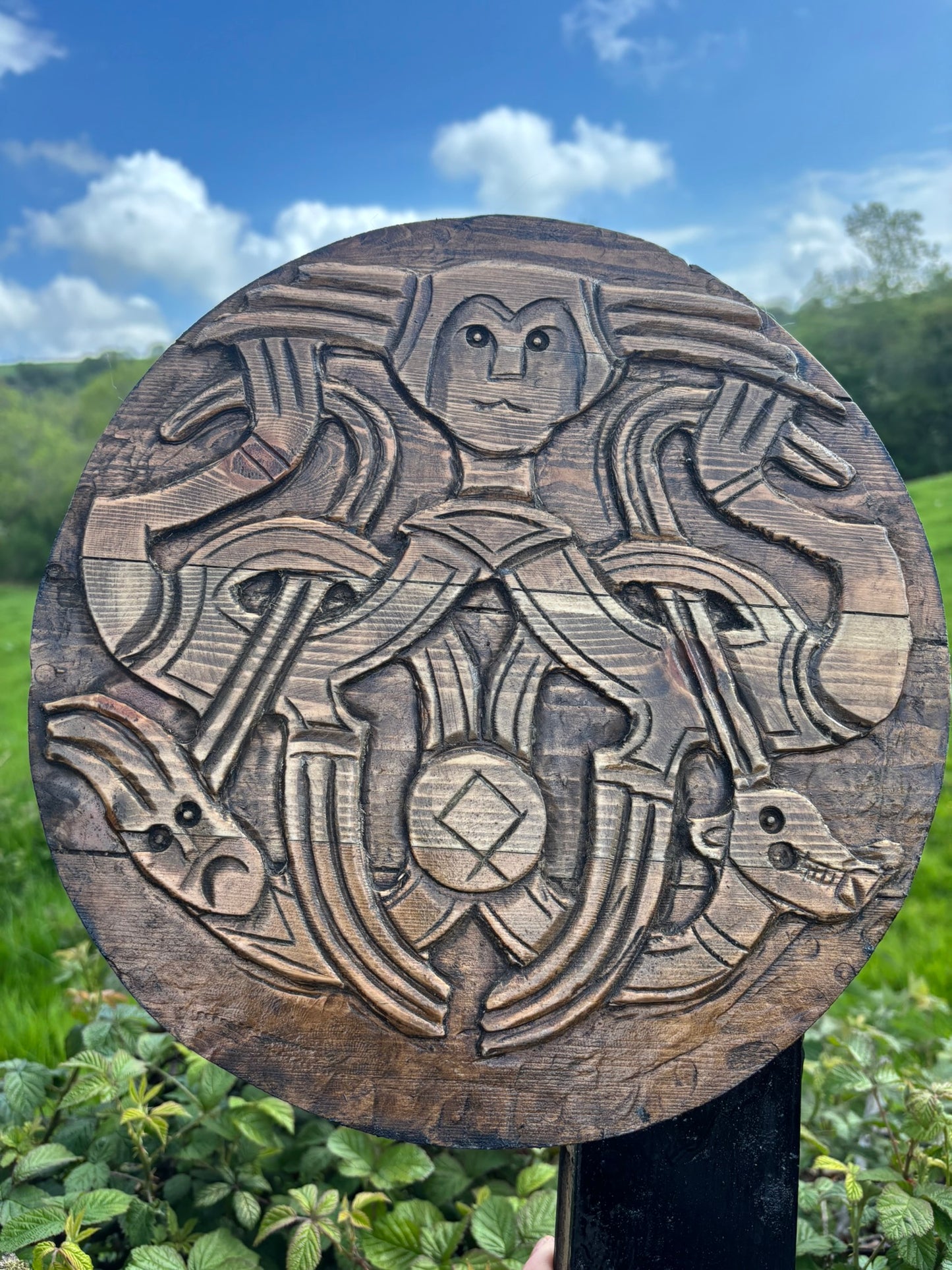 A round, hand-carved wooden plaque featuring intricate Norse-style animal and human motifs, mounted on a black post against a green outdoor background with foliage and a blue sky.