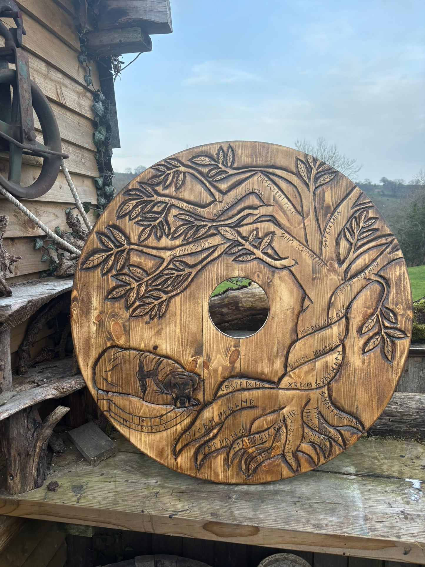 A large circular wooden spool carved with a tree of life design, featuring engraved names and branches, displayed outdoors on a rustic wooden deck next to a wooden structure.
