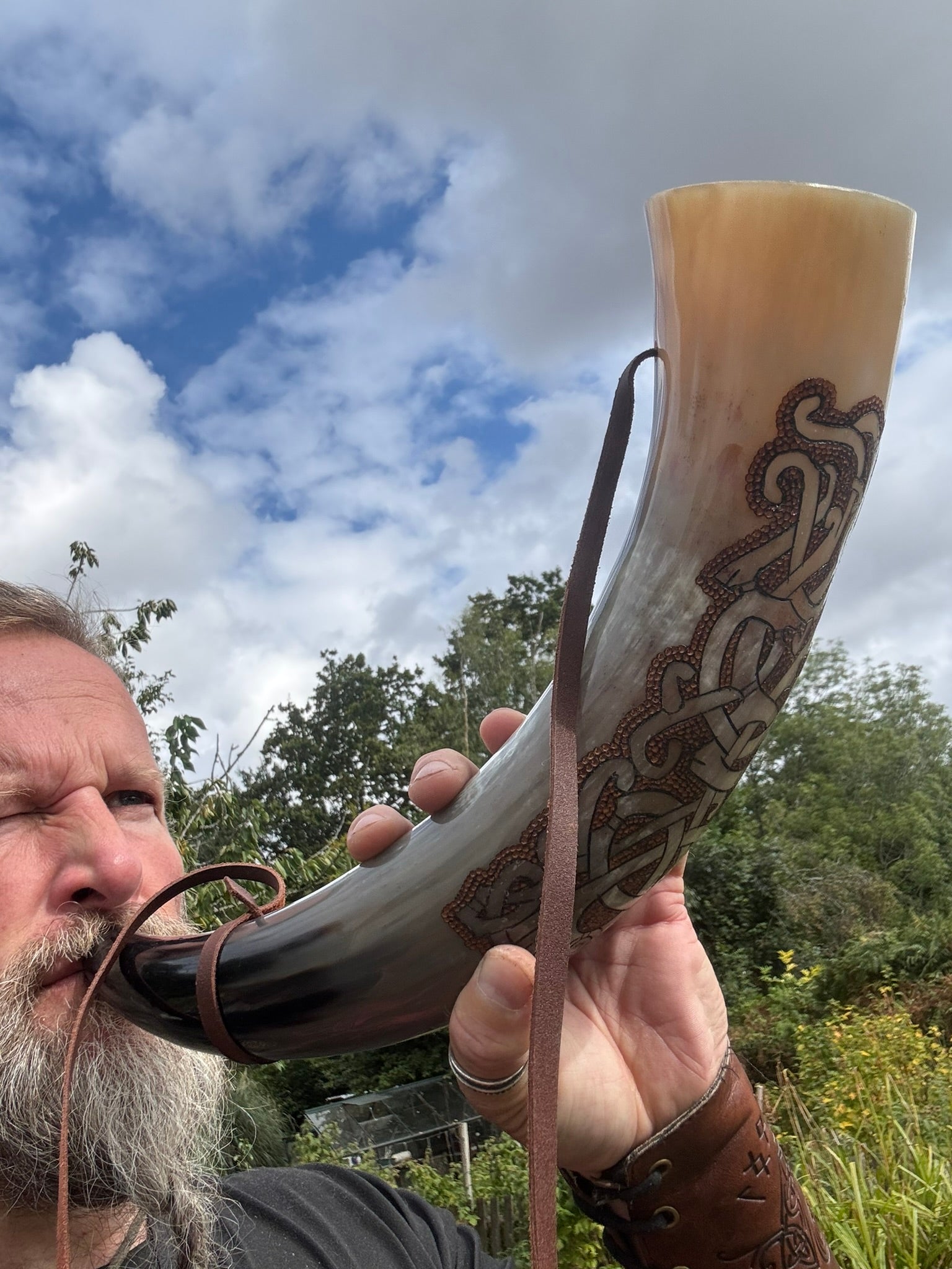 A man outdoors blowing into a large natural horn shofar with brown leather straps and carved in the Mammen Viking Style detailing, against a cloudy blue sky and green trees.