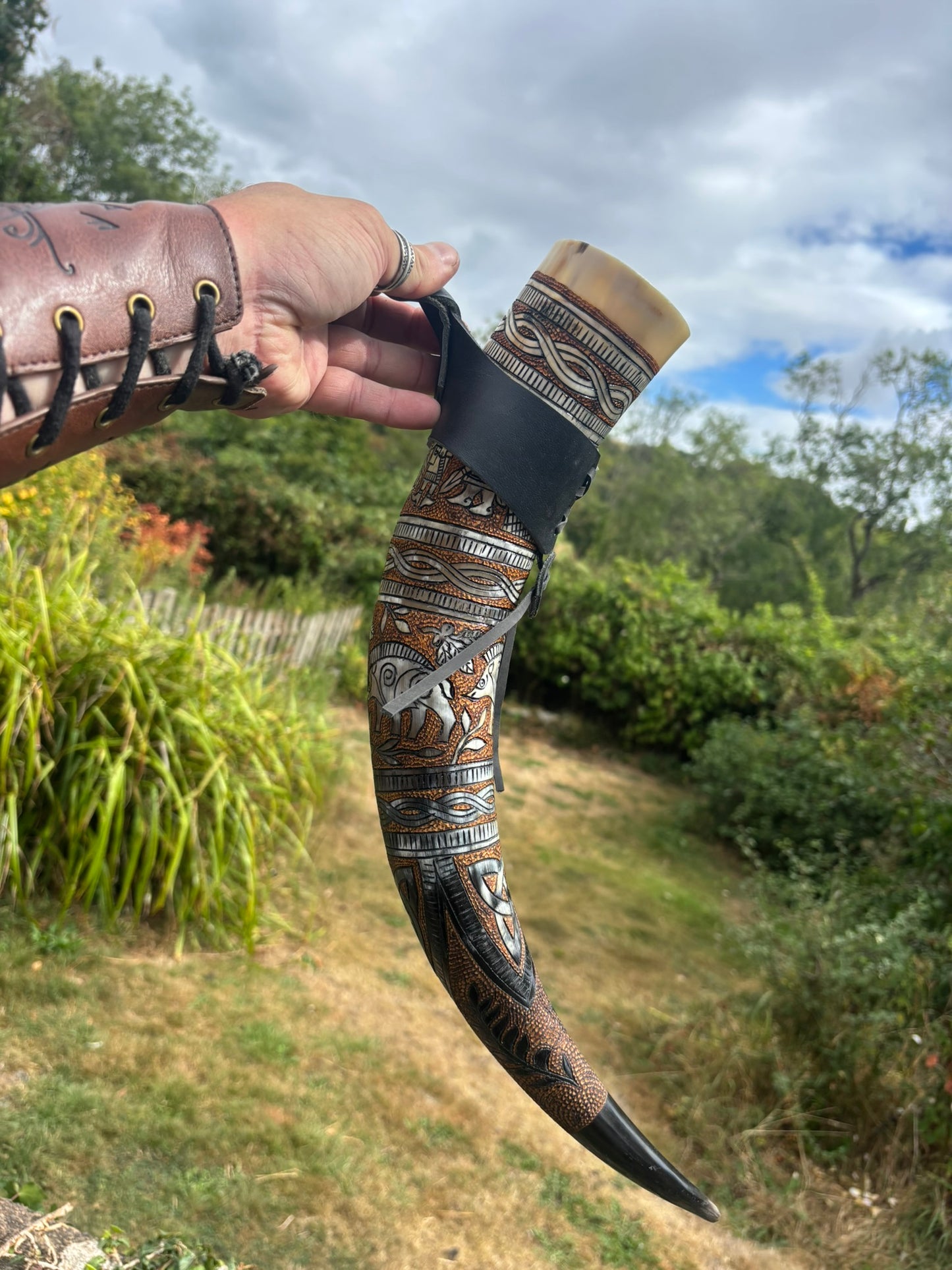 A hand holds a decorated curved horn or tusk-style object outdoors, with a black leather strap around the upper section. The item features intricate engraved geometric and animal motifs in tan, brown, black, and white, and the background shows grass, shrubs, trees, and a cloudy sky.