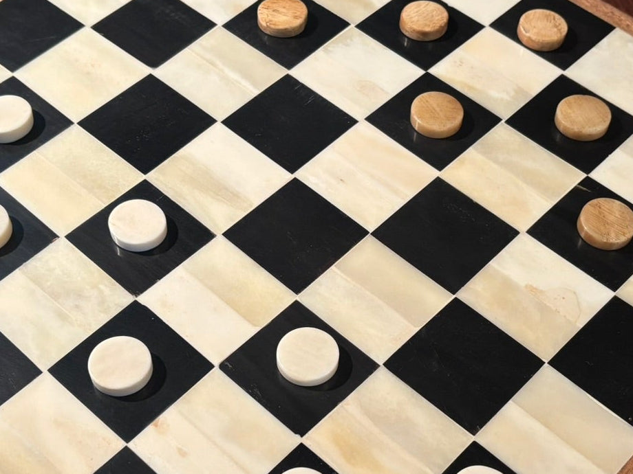 A square checkers board made from black and cream-colored bone, with round checkers, displayed on a dark wooden table.