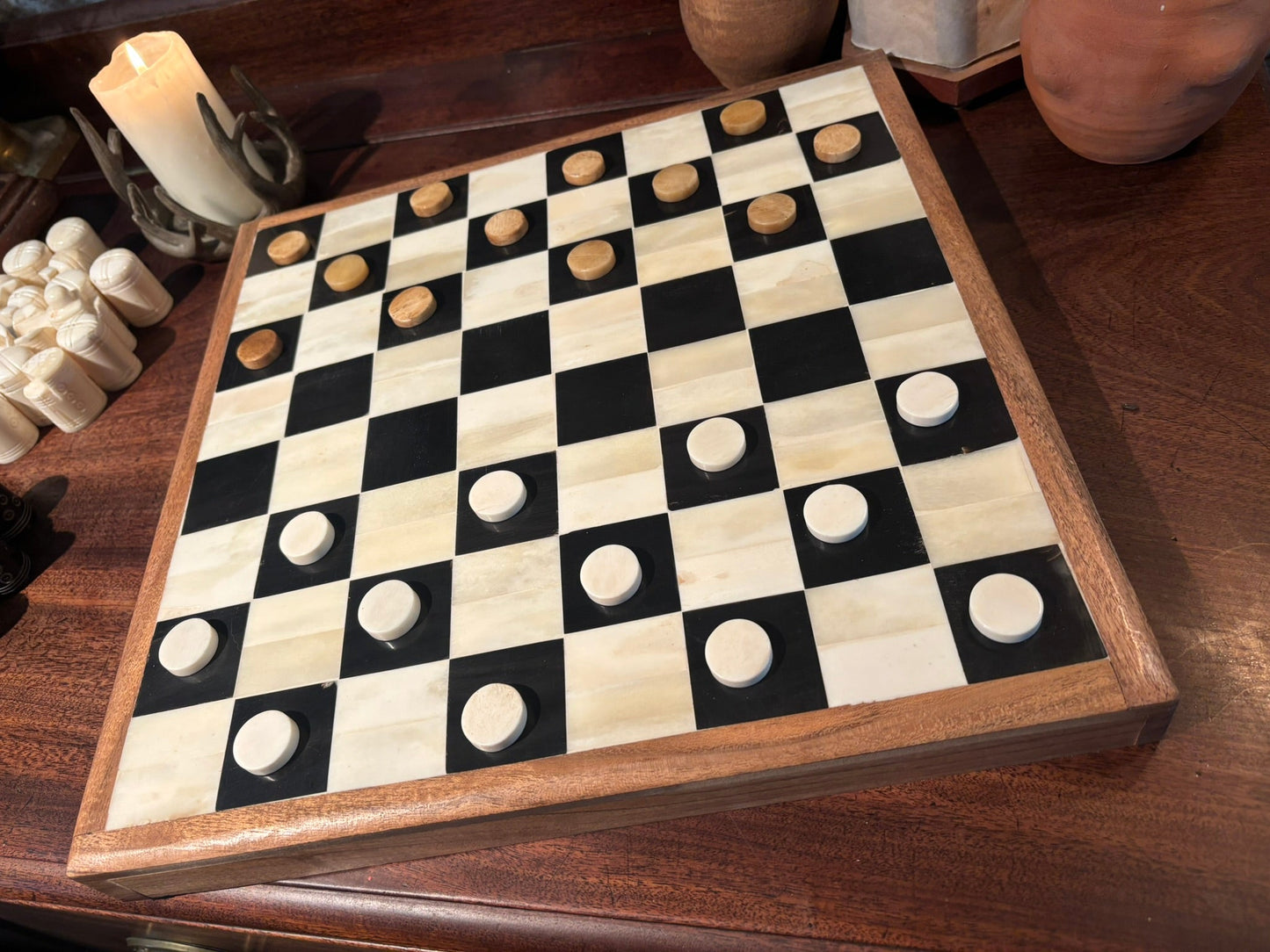 A square Bone and Horn checkers board with black and cream checkered squares, bone pieces, and white cylindrical pieces, photographed on a dark wood tabletop with candles in the background.