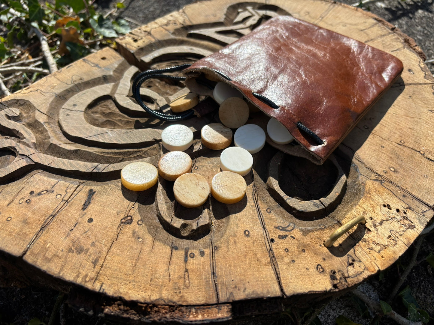 A brown leather pouch with a black cord lies open on a carved wooden surface outdoors, with several round bone checkers game pieces spilled out beside it.