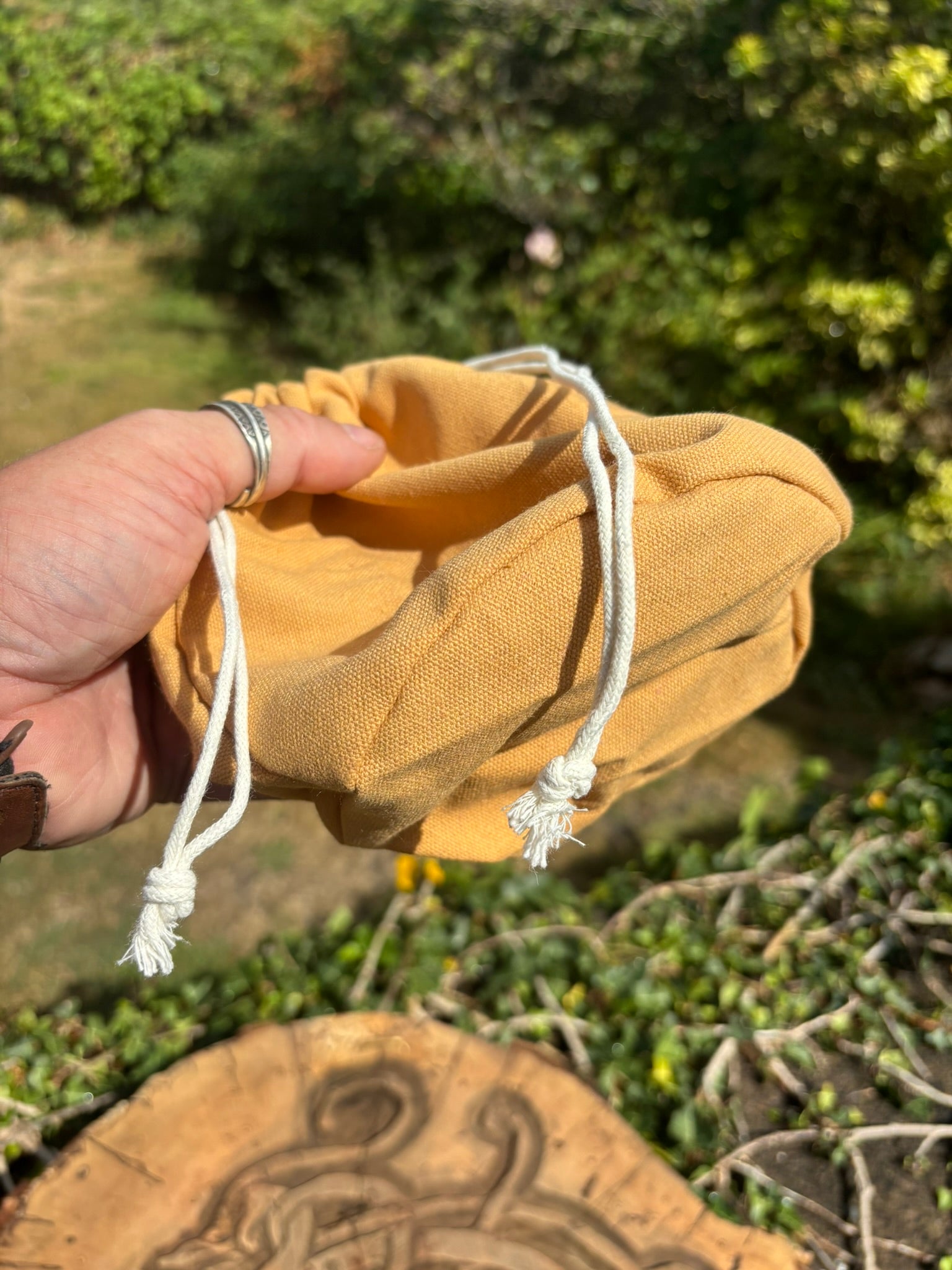 A bushcrafter holding a small mustard yellow fabric pouch with white drawstrings, set against a blurred outdoor garden background.