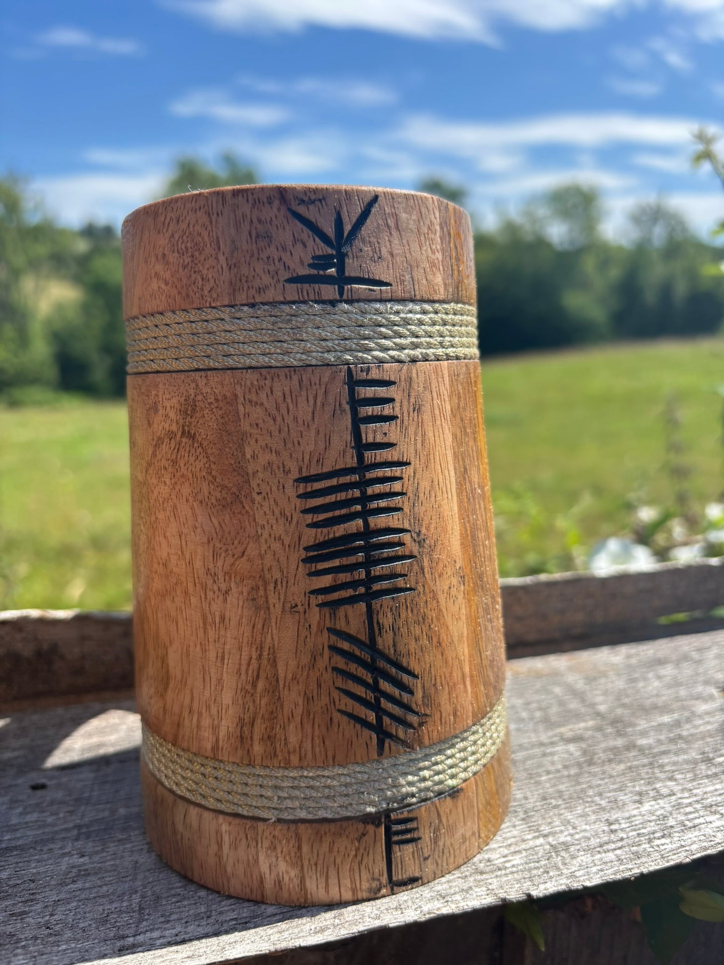 A cylindrical wooden box with engraved black symbols and two wrapped bands of natural rope, shown outdoors on a weathered wooden surface with a blurred green field and blue sky in the background.