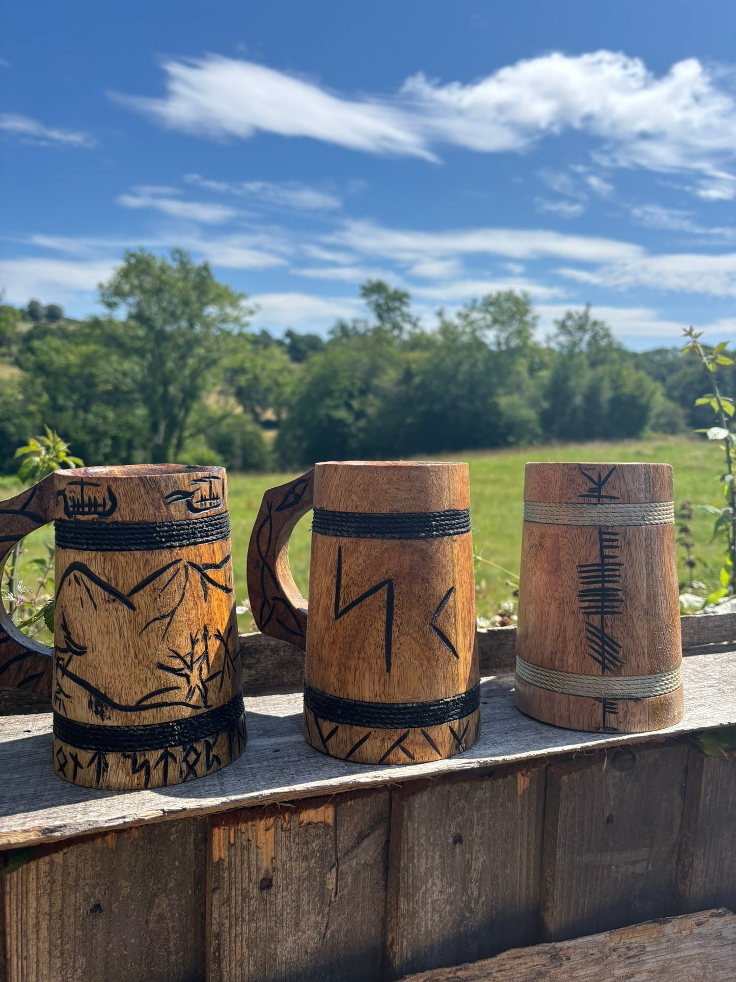 Three handcrafted wooden beer mugs with Norse-style carvings and black rope bands, displayed outdoors on a wooden railing with a green field and blue sky in the background.