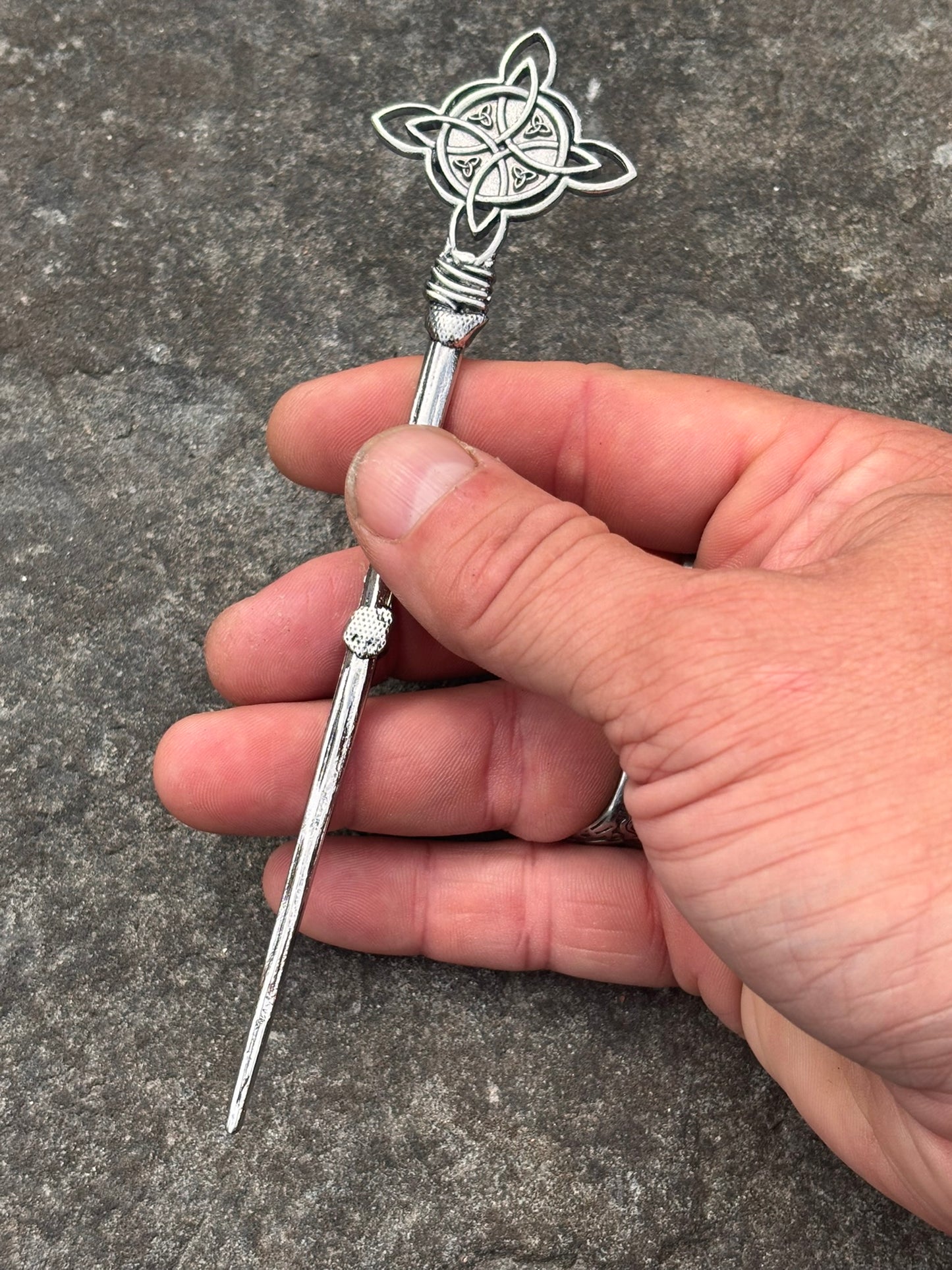 A hand holding a Norse silver-toned metal hair  pin with a Celtic knotwork design on the top and a small clear stone on the shaft, set against a gray textured stone background.