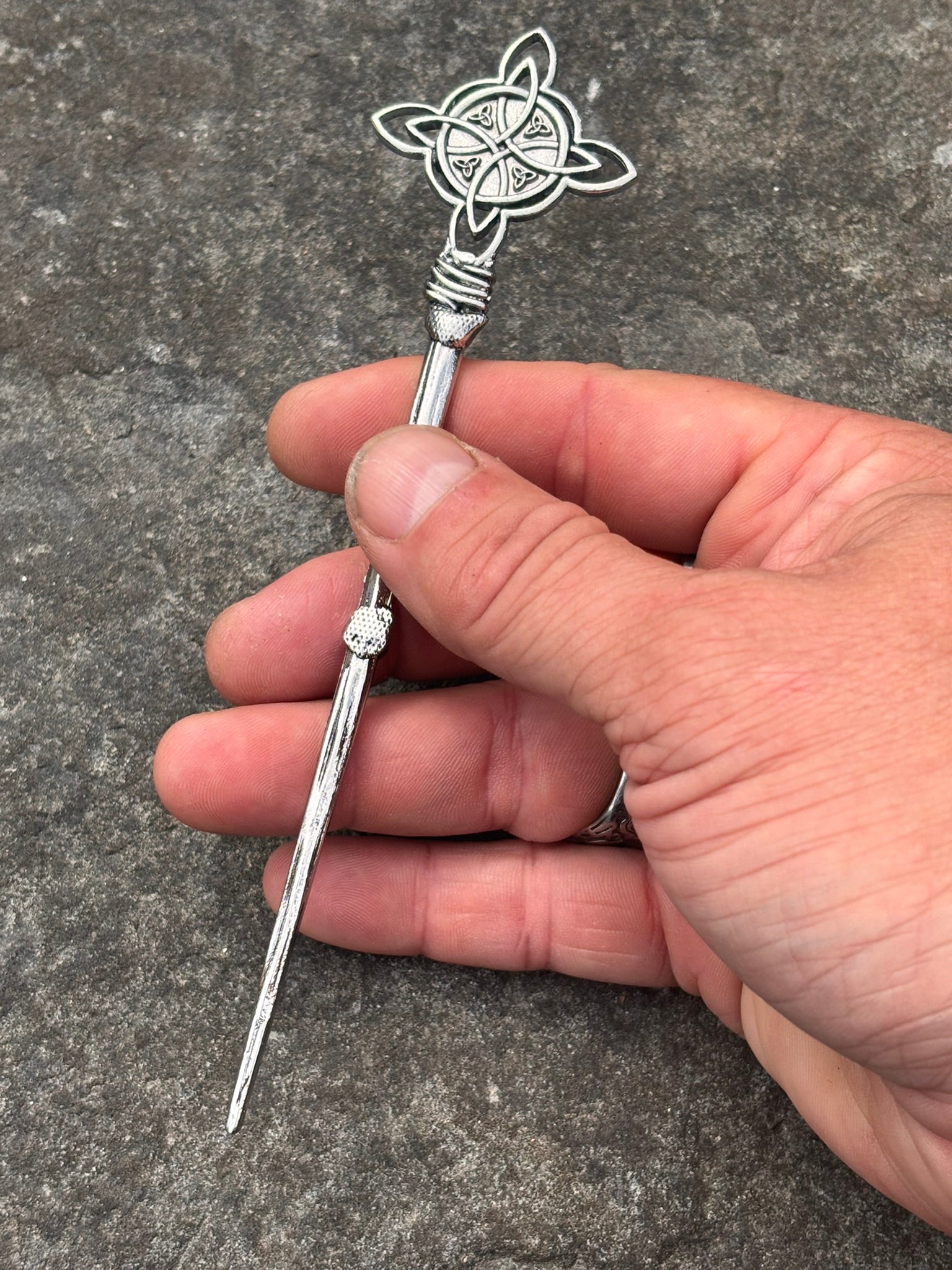 A hand holding a Norse silver-toned metal hair  pin with a Celtic knotwork design on the top and a small clear stone on the shaft, set against a gray textured stone background.