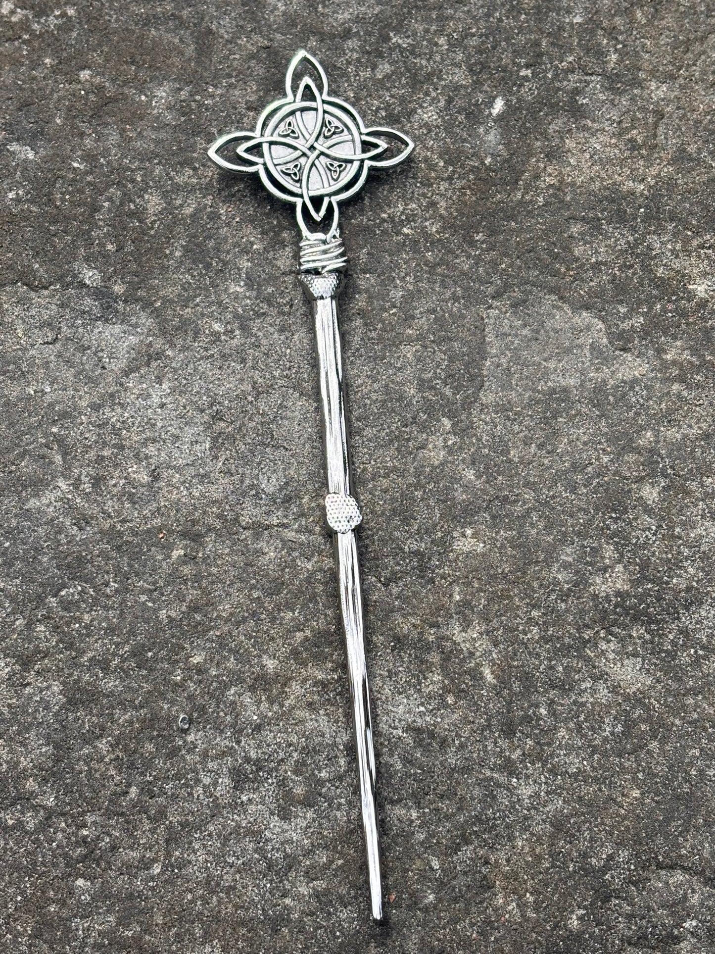 A silver-toned metal hair stick with a Celtic knotwork design on the top and a twisted stem, displayed on a textured gray stone surface.