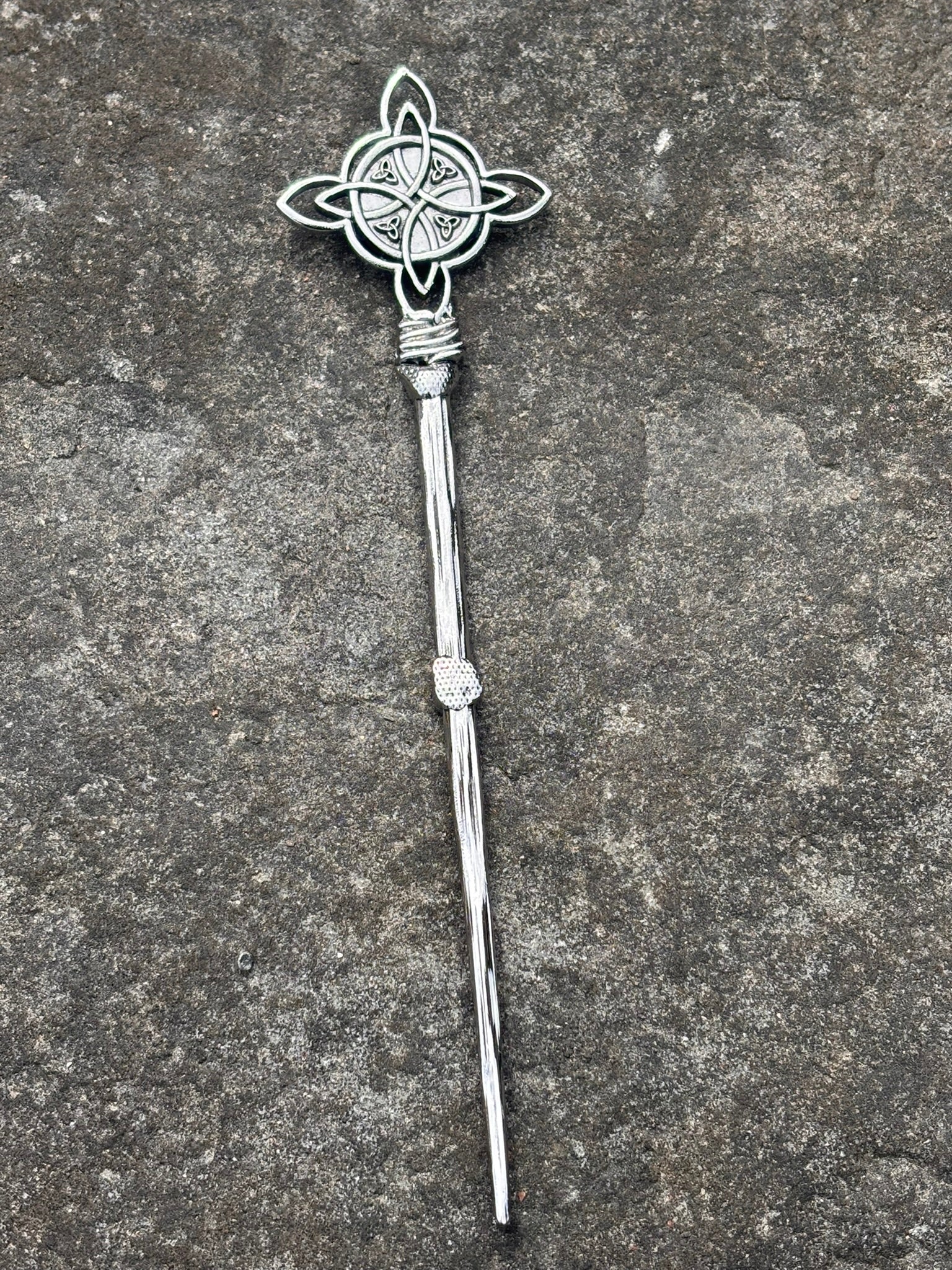 A silver-toned metal hair stick with a Celtic knotwork design on the top and a twisted stem, displayed on a textured gray stone surface.