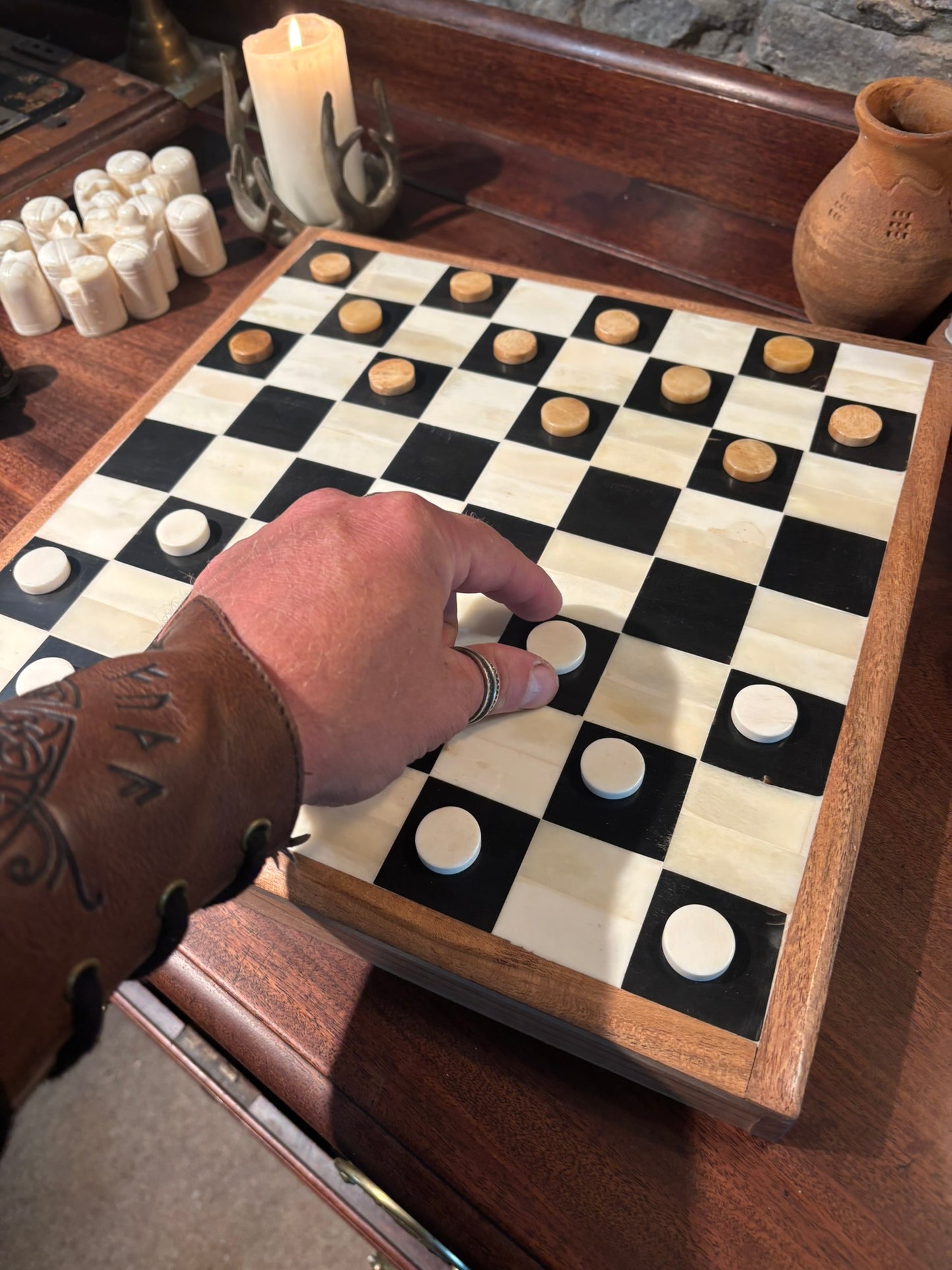 A Horn and bone checkers board with black and cream bone checkers, shown on a wooden table with a person's hand placing a white checker. Decorative candles and a small vase are visible in the background.