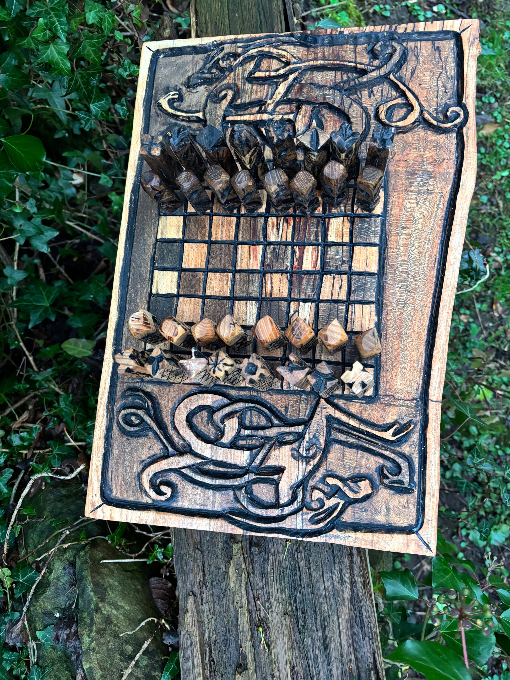 A handcrafted wooden board game set with carved Celtic knot designs and wooden chess pieces, displayed outdoors on a weathered wooden post with green foliage in the background.