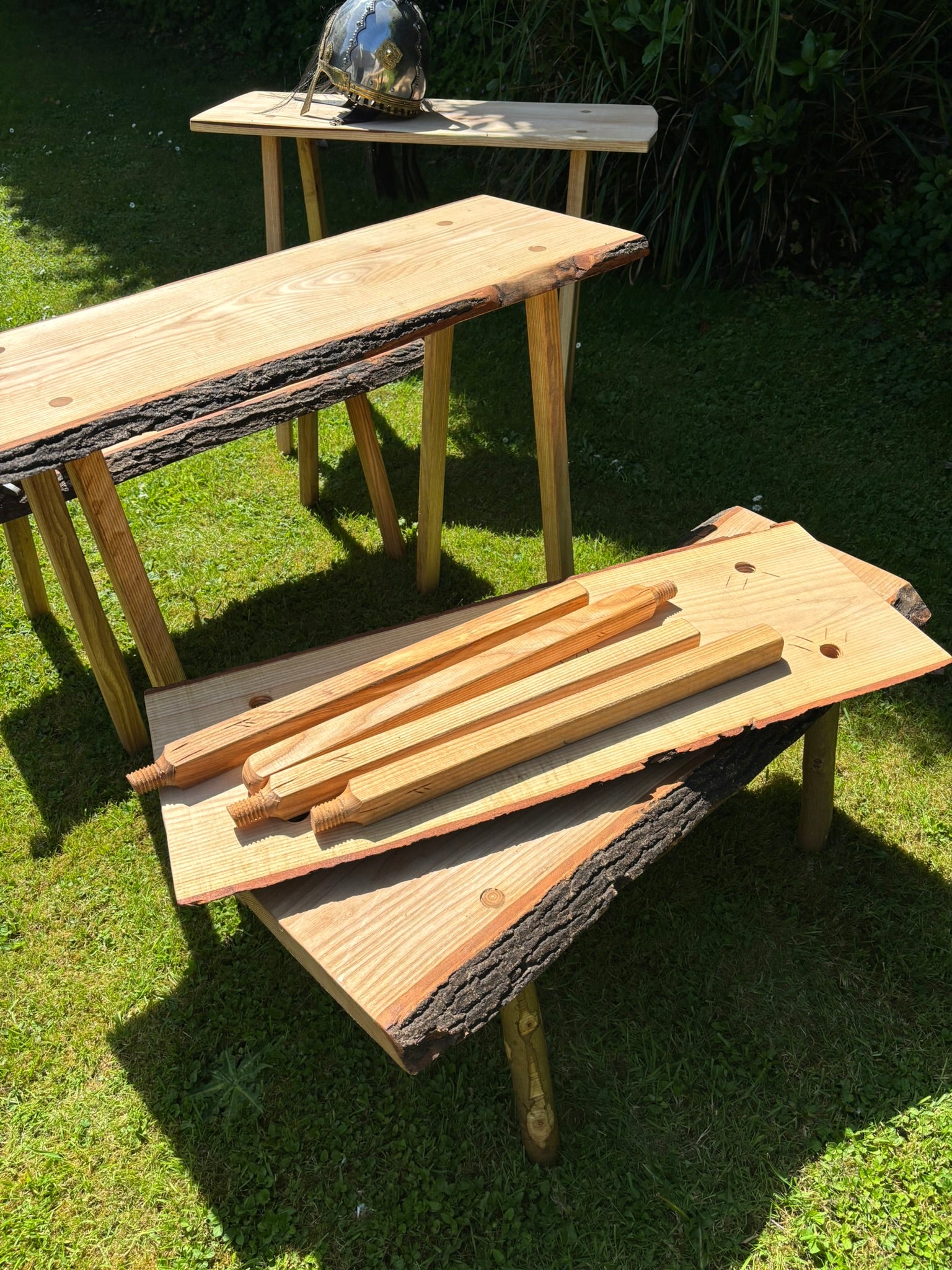 Three rustic live-edge wood tables with natural bark trim are arranged on a grassy lawn outdoors. The tables have light tan wood tops and legs, with one table holding a dark metal bowl and the others displaying wood pieces on top.