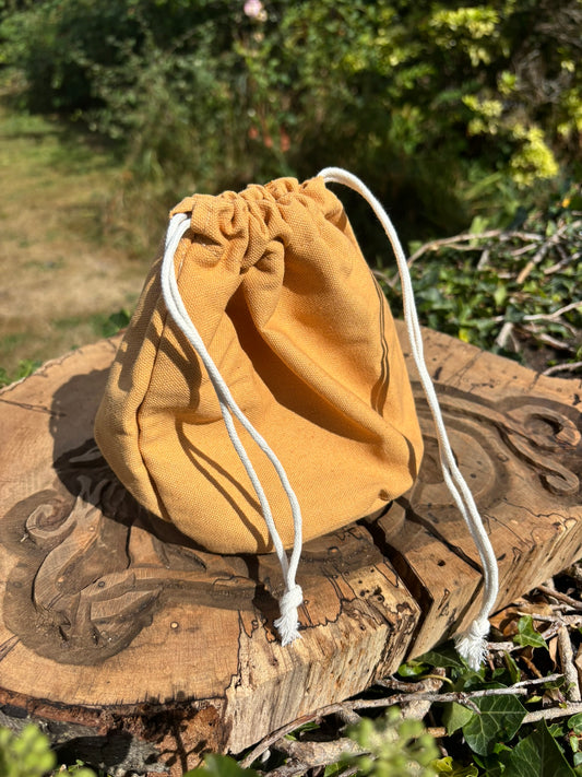 A mustard yellow drawstring pouch bag made of soft fabric, resting on a rustic carved wooden log outdoors with green foliage in the background.