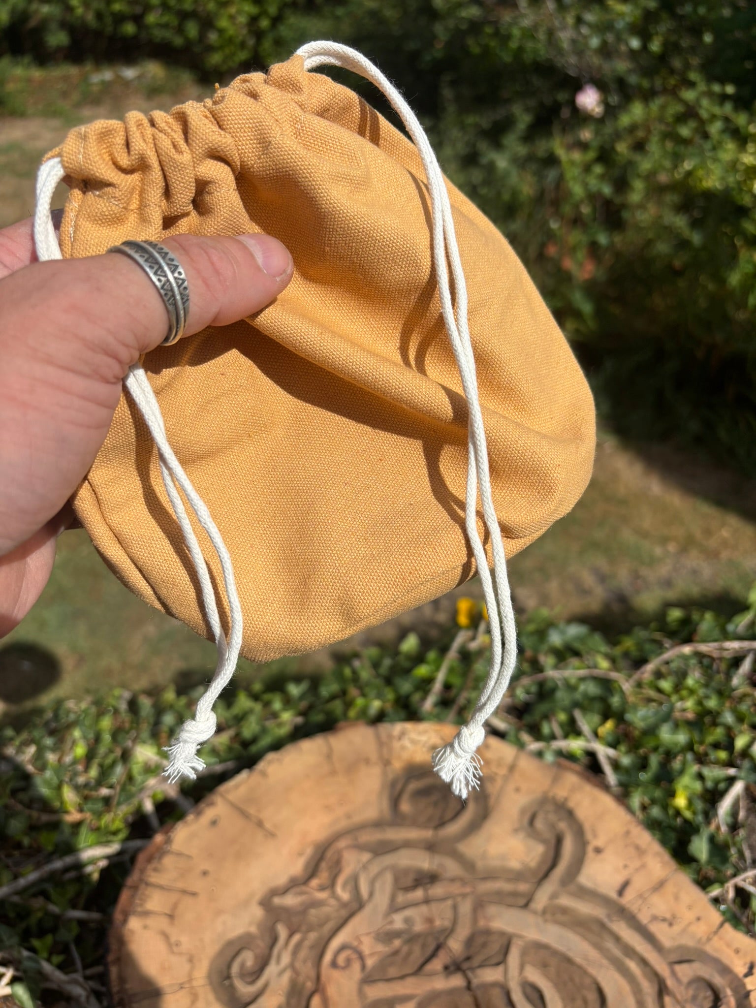 A mustard yellow fabric drawstring pouch held by a hand outdoors, with green foliage and a carved wooden slice in the background.