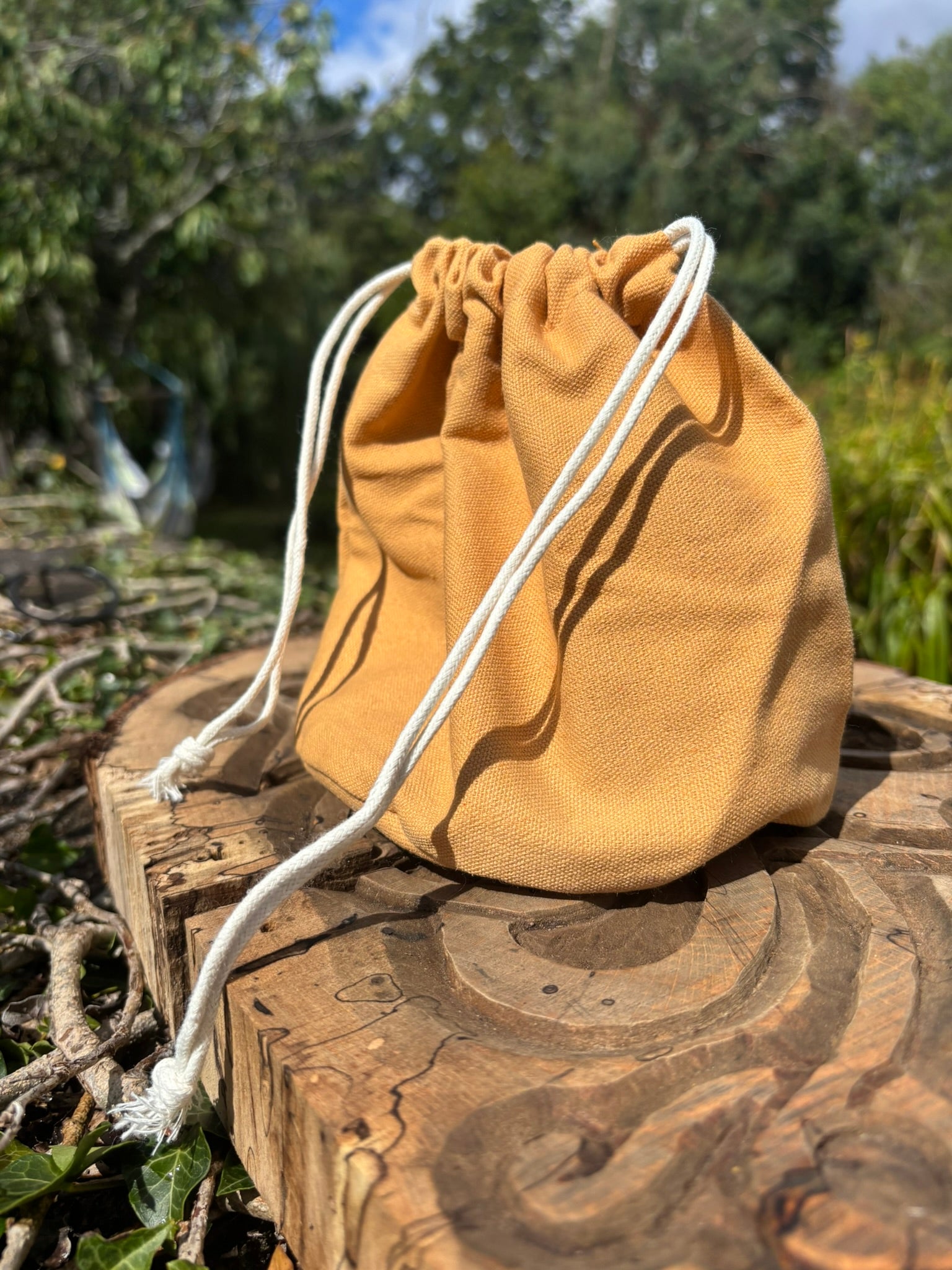 A small mustard yellow canvas drawstring pouch with white cords, resting on a rustic wooden log outdoors with trees and foliage in the background.