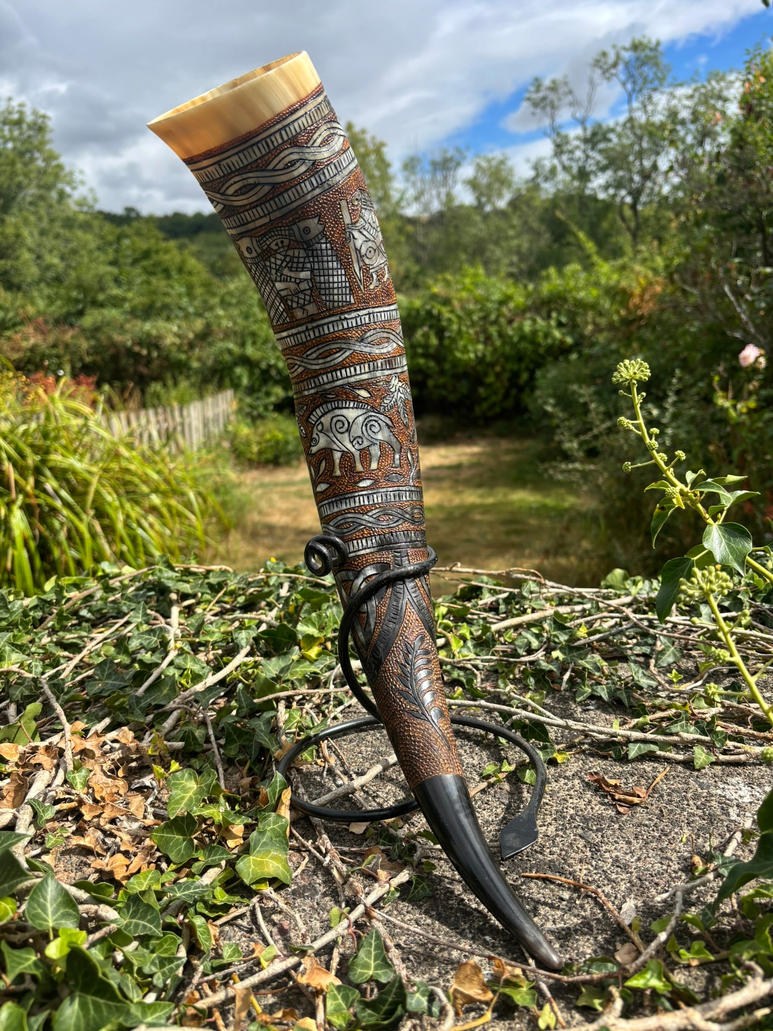 A carved horn showing the Poetic Edda with intricate silver and brown patterns, displayed outdoors on a stone surface with a garden background.
