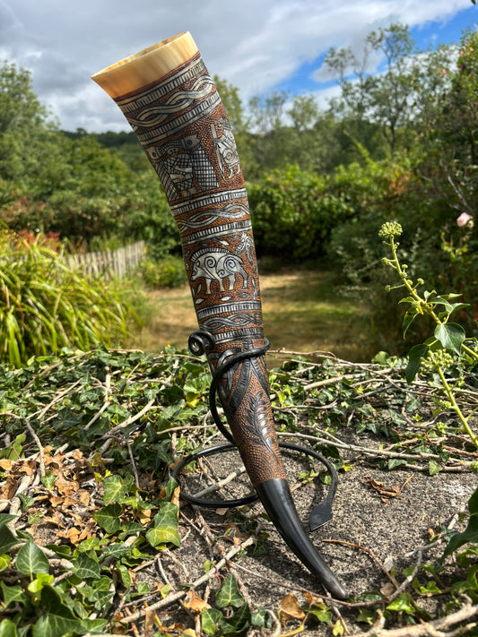 A carved horn showing the Poetic Edda with intricate silver and brown patterns, displayed outdoors on a stone surface with a garden background.
