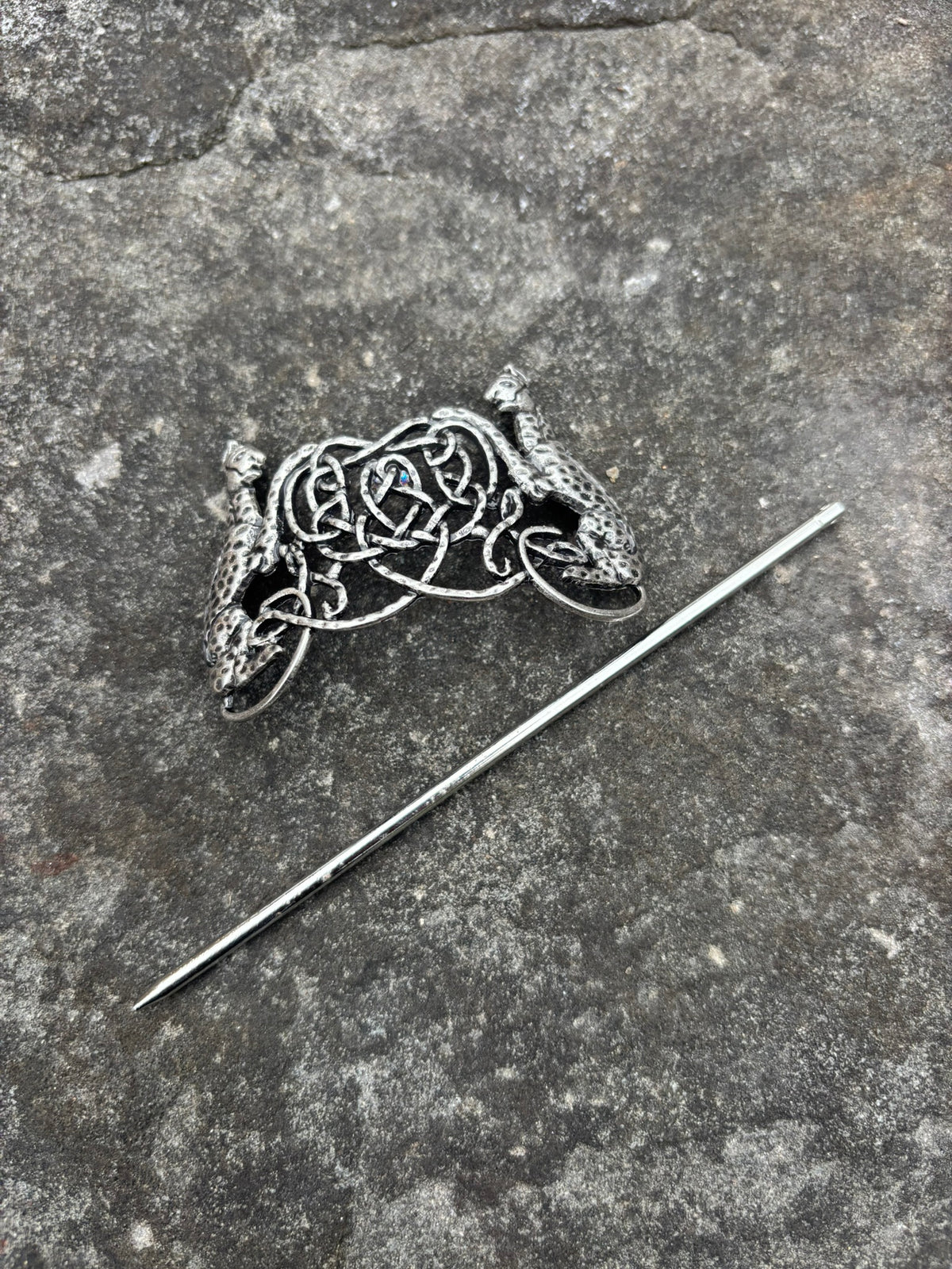 A silver-toned Celtic knotwork hair pin with dragon motifs on either side, displayed diagonally on a textured gray stone surface next to its matching straight pin.