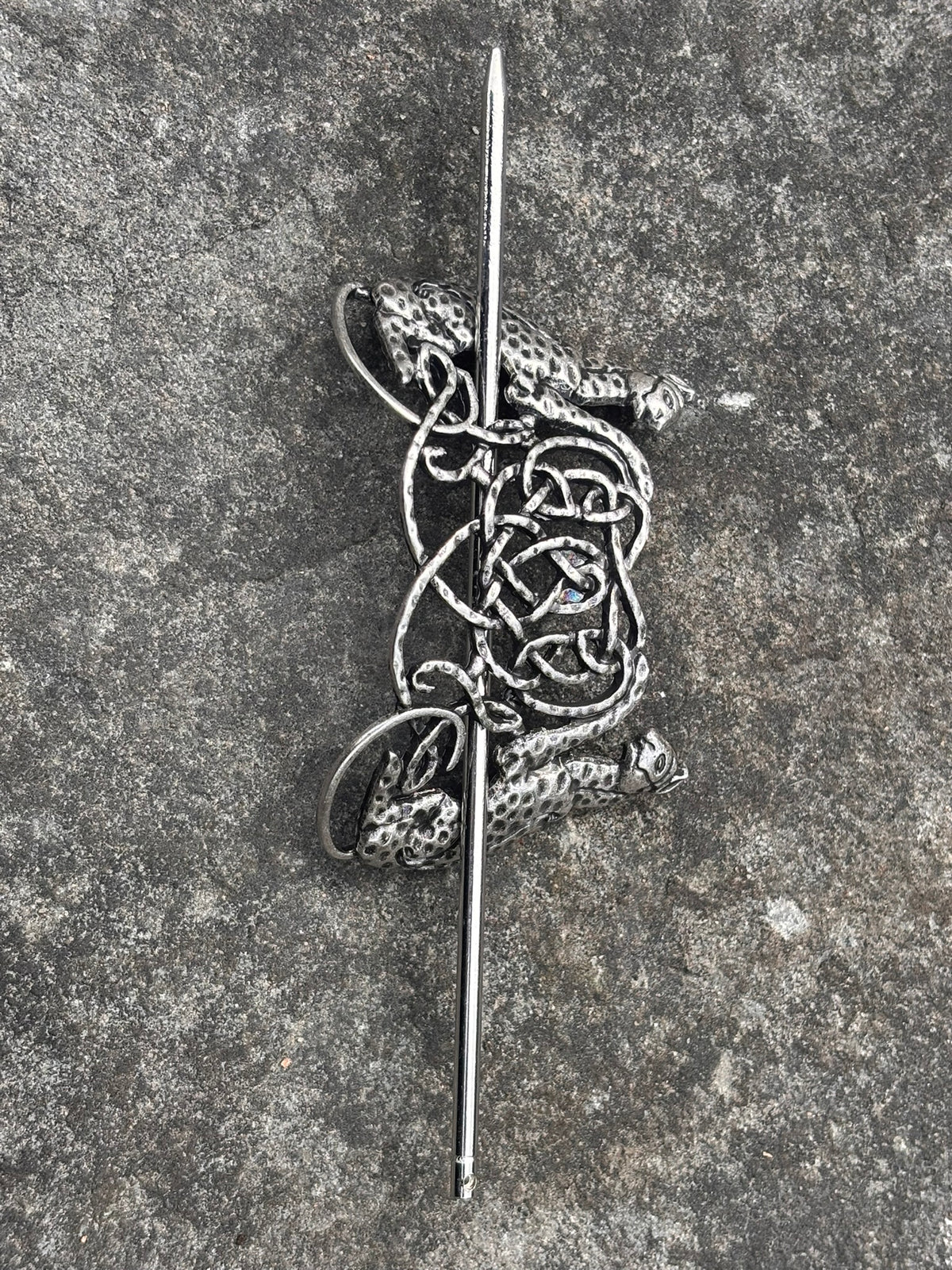 A silver-toned Celtic knotwork brooch featuring a stylized cat motif, photographed flat on a textured gray stone surface.