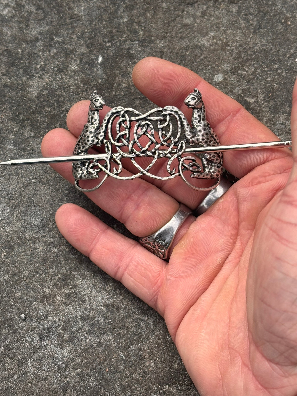 A silver-toned Celtic-style brooch with Freya's two cats and an interlaced knotwork heart design, held in a person's hand against a gray textured background.