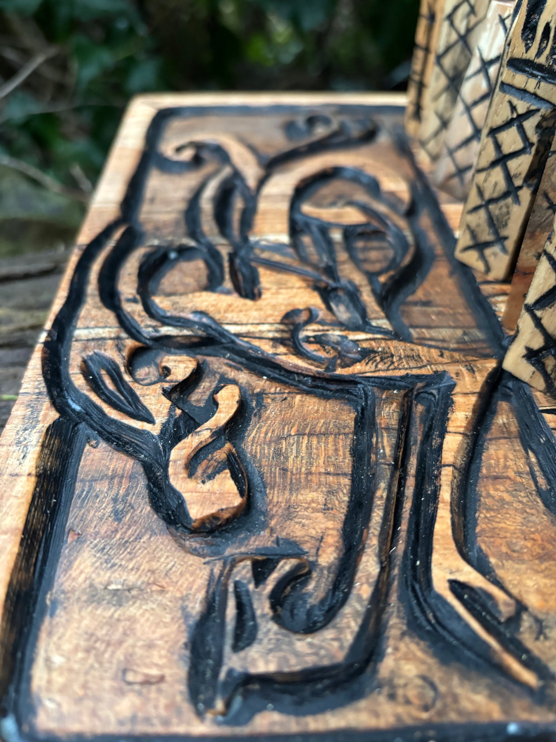 A close-up of a hand-carved wooden  chess board featuring a stylized deer head with antlers in dark burnished contrast against a warm natural wood grain, with a blurred outdoor background.