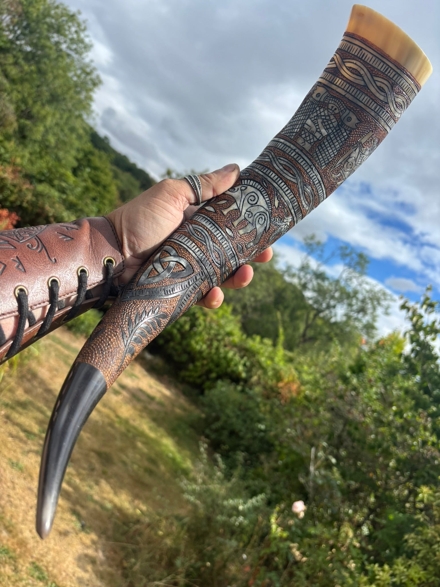 A hand holds a carved horn or antler object outdoors, decorated with dark geometric and tribal-style patterns. The item has a light tan end, a dark black tip, and a brown leather sleeve with lace holes on the side.