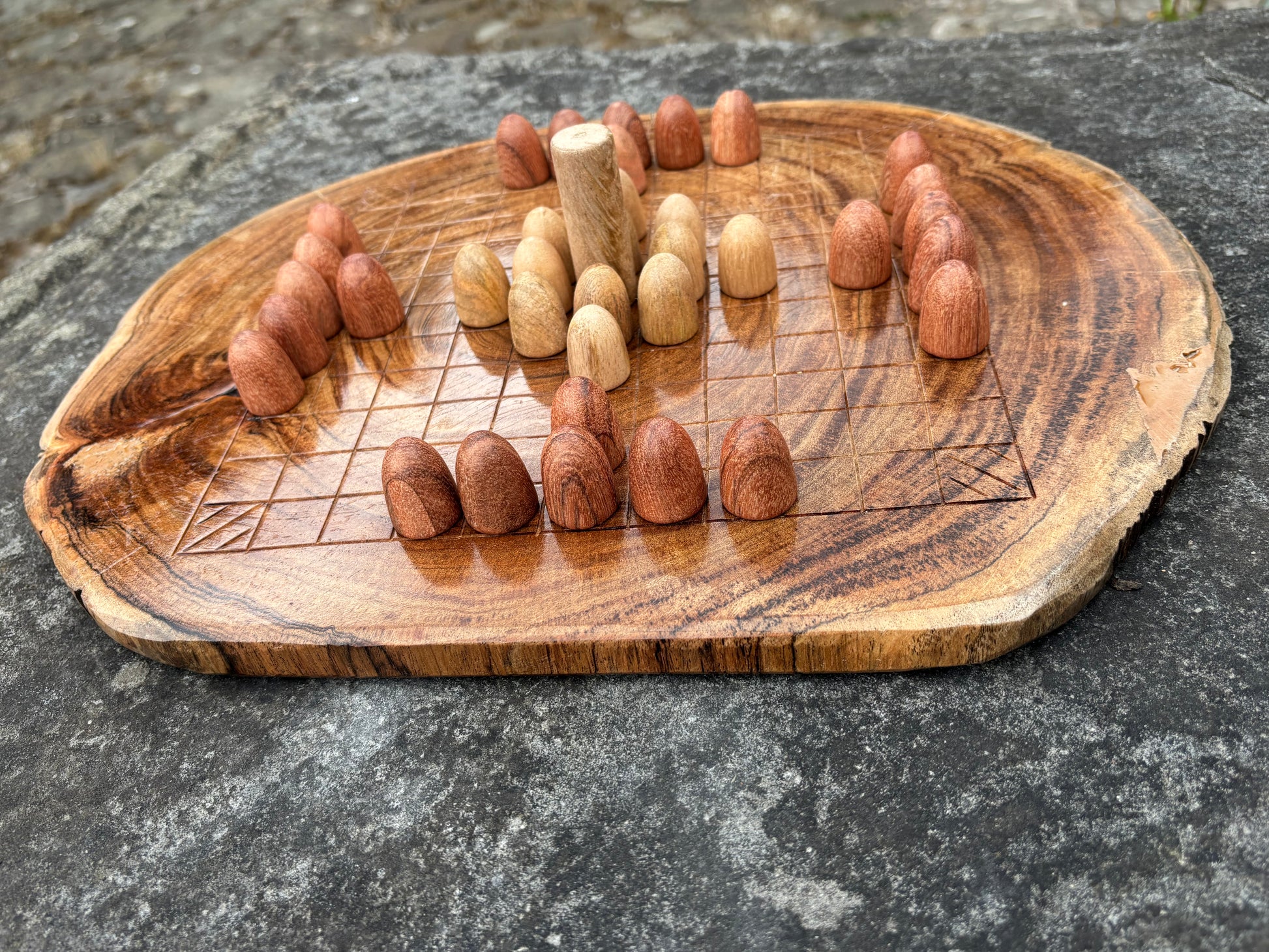 A rustic wooden game board made from a natural tree slice, featuring carved wooden pieces and a central peg, set on a gray stone surface.