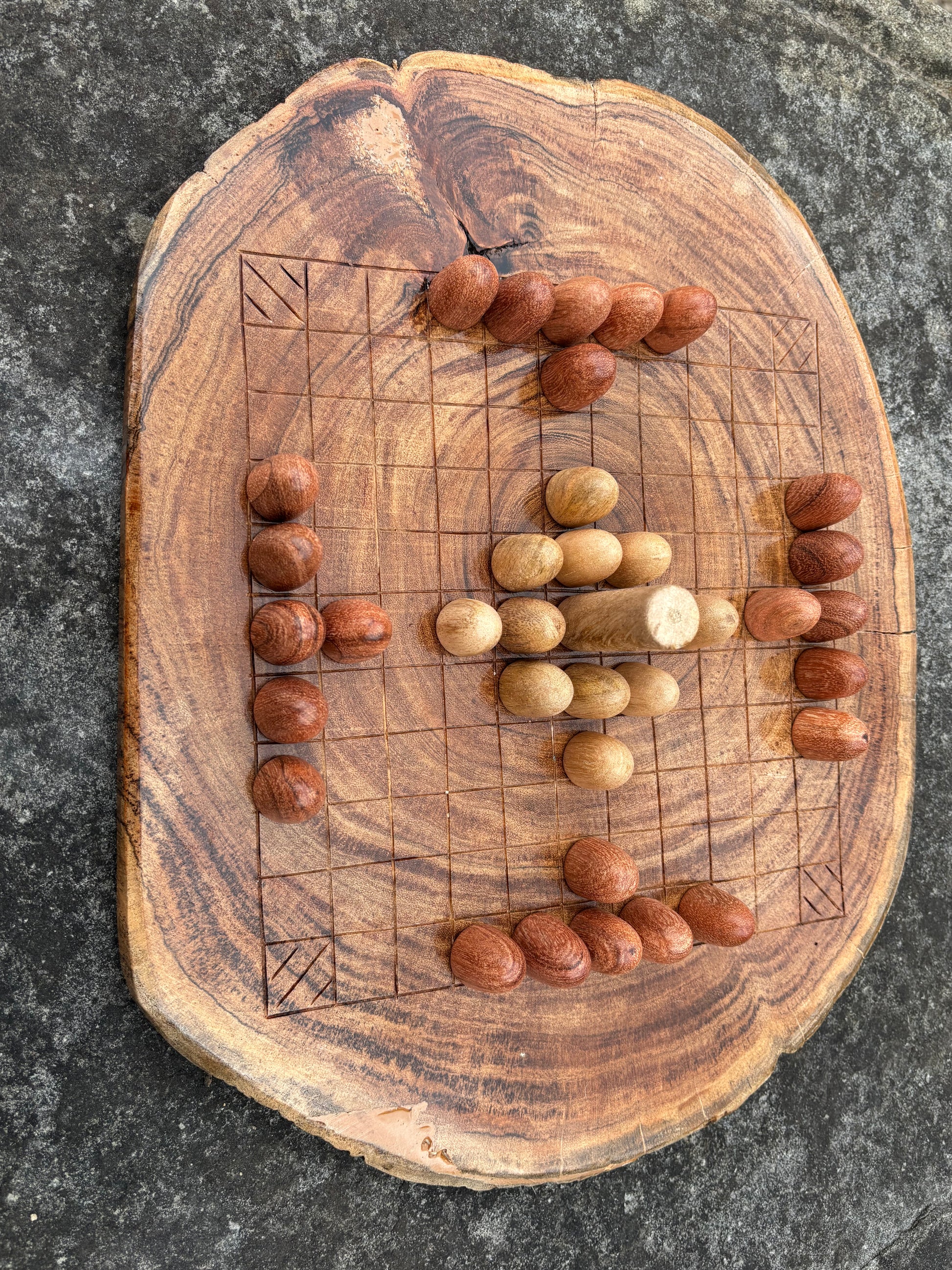 A top-down view of a rustic wooden game board made from a natural tree slice, featuring a grid and small round wooden pieces, placed on a dark textured stone surface.