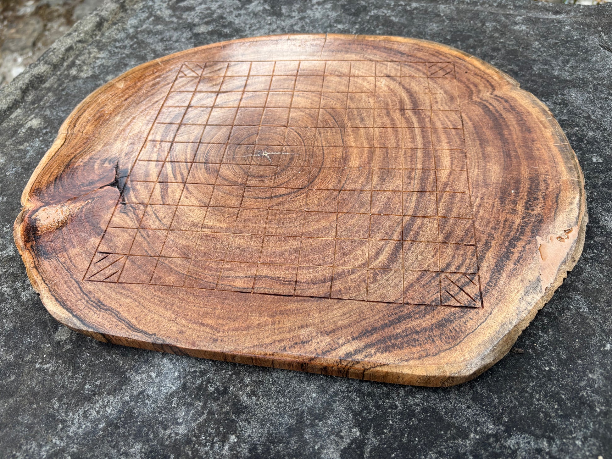 A rustic wooden chess board made from a natural tree slice, featuring engraved grid lines and corner markings, placed on a dark textured stone surface.