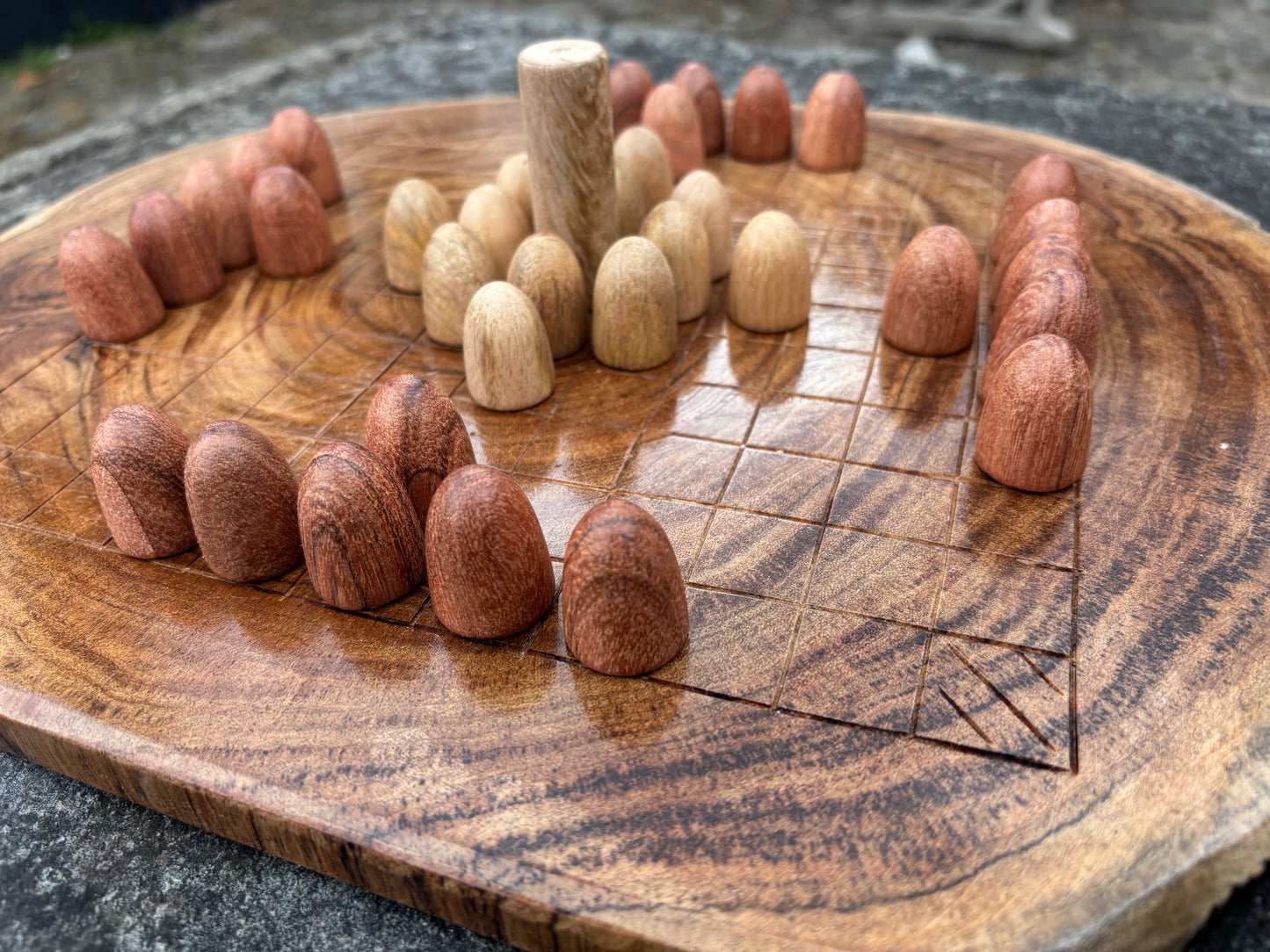 A close-up of a round wooden board game set with small wooden pieces arranged on a carved wooden board outdoors.