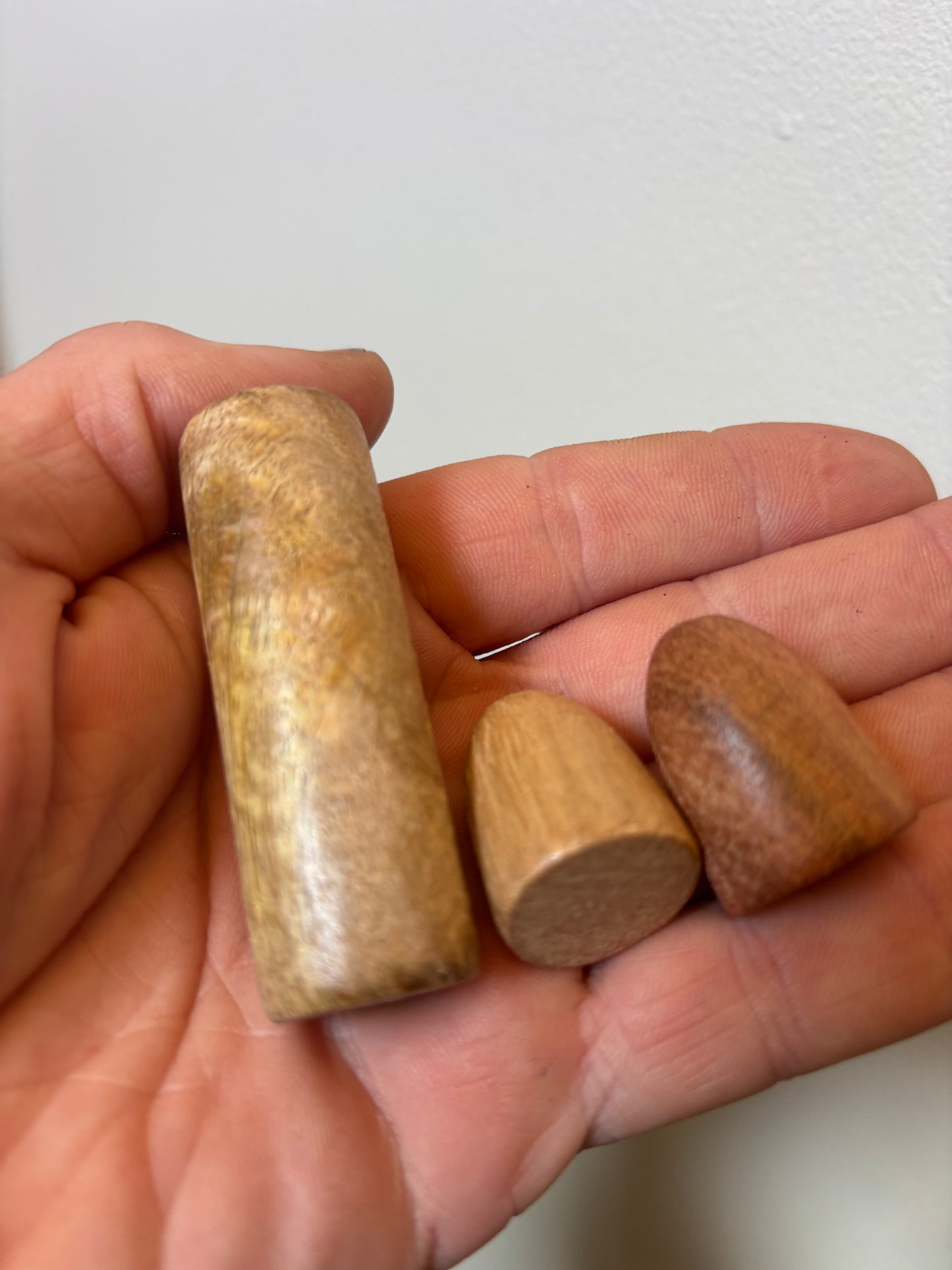 Hnefatafl Set — Plain Wood Three small wooden pieces held in a person's hand against a plain light wall. The set includes one tall cylindrical piece and two smaller rounded pieces in different wood tones.