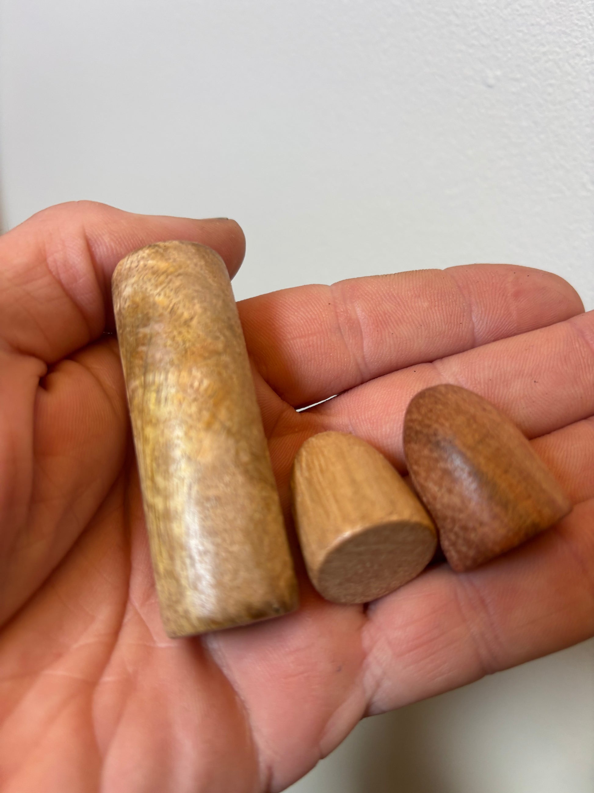 Hnefatafl Set — Plain Wood Three small wooden pieces held in a person's hand against a plain light wall. The set includes one tall cylindrical piece and two smaller rounded pieces in different wood tones.