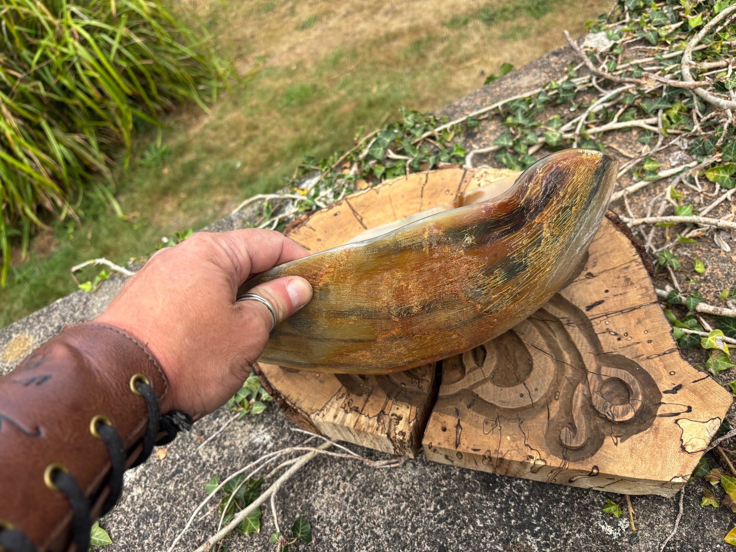 A Viking holds a carved natural horn bowl over a carved wooden block outdoors on grass and stone.