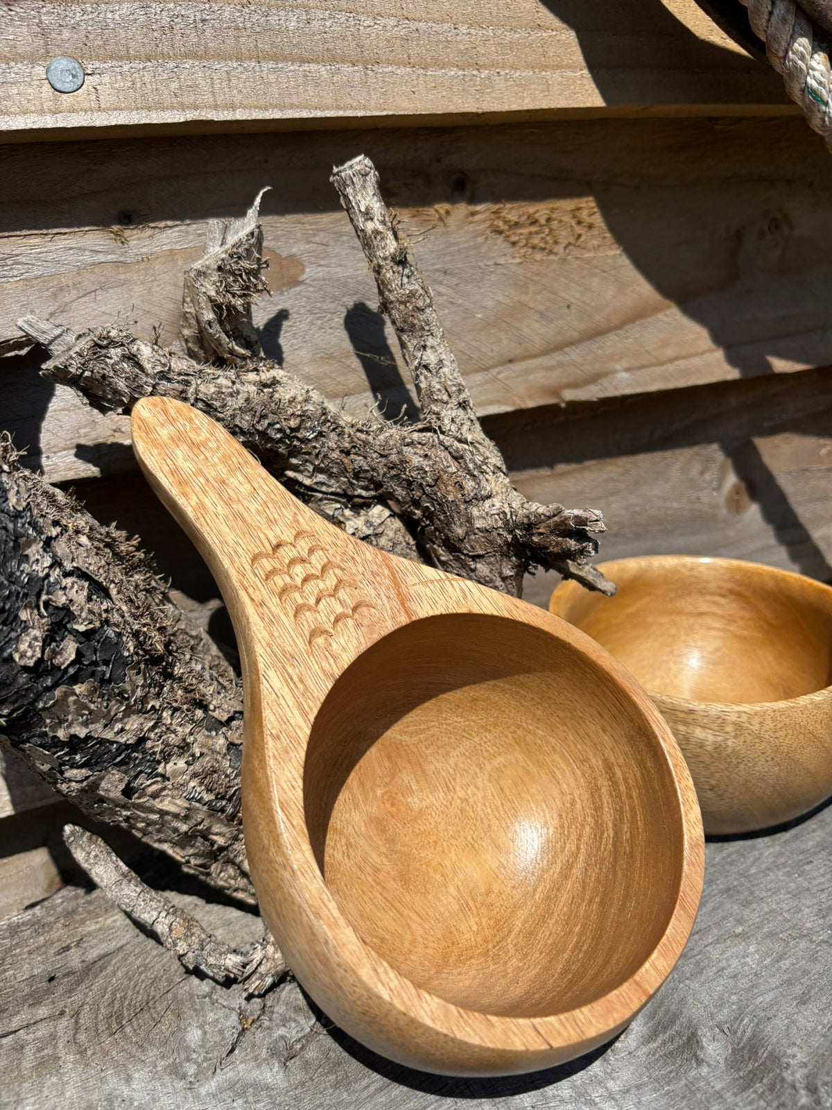A handcrafted wooden Kuksa with a carved handle, shown alongside a matching wooden bowl on a rustic wood background.