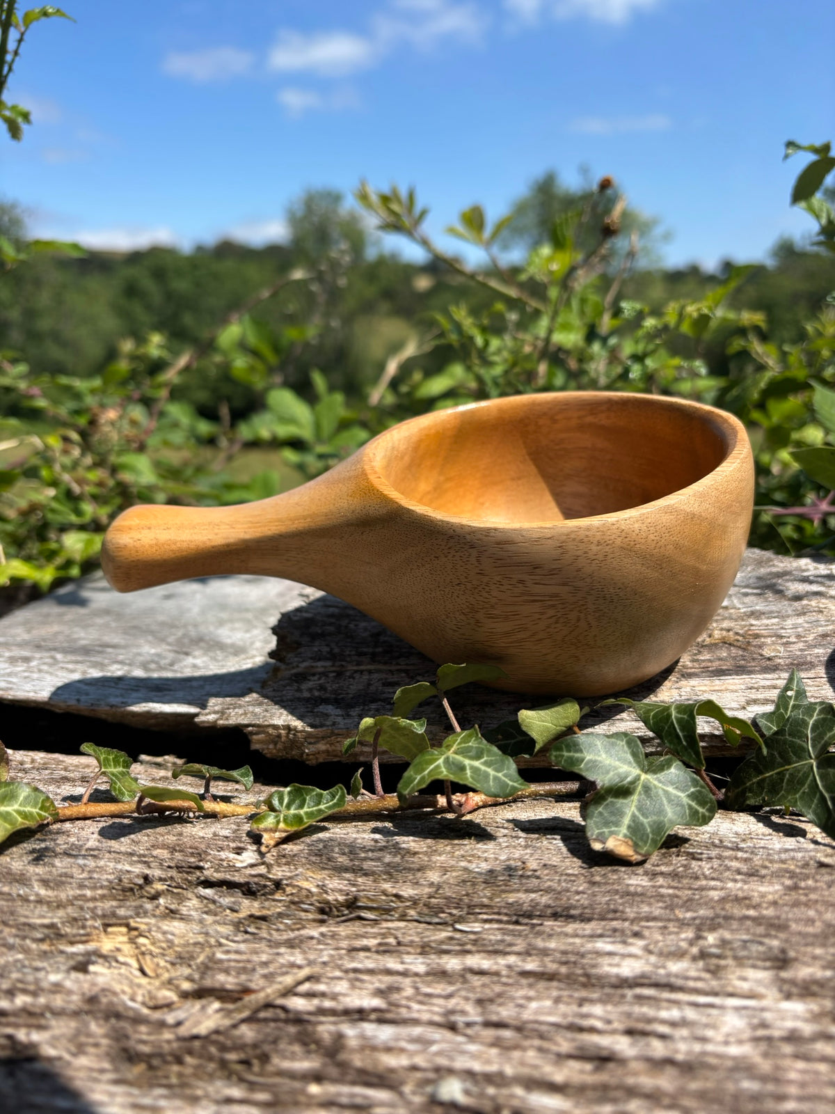 A Kuksa with a handle, resting on a weathered log outdoors with ivy leaves and a blurred green garden background under a blue sky.