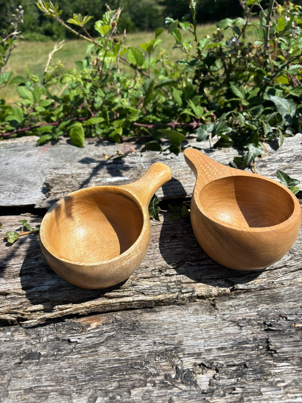 Two small handcrafted wooden Kuksas with integrated handles, placed on a weathered wooden surface outdoors with green foliage in the background.