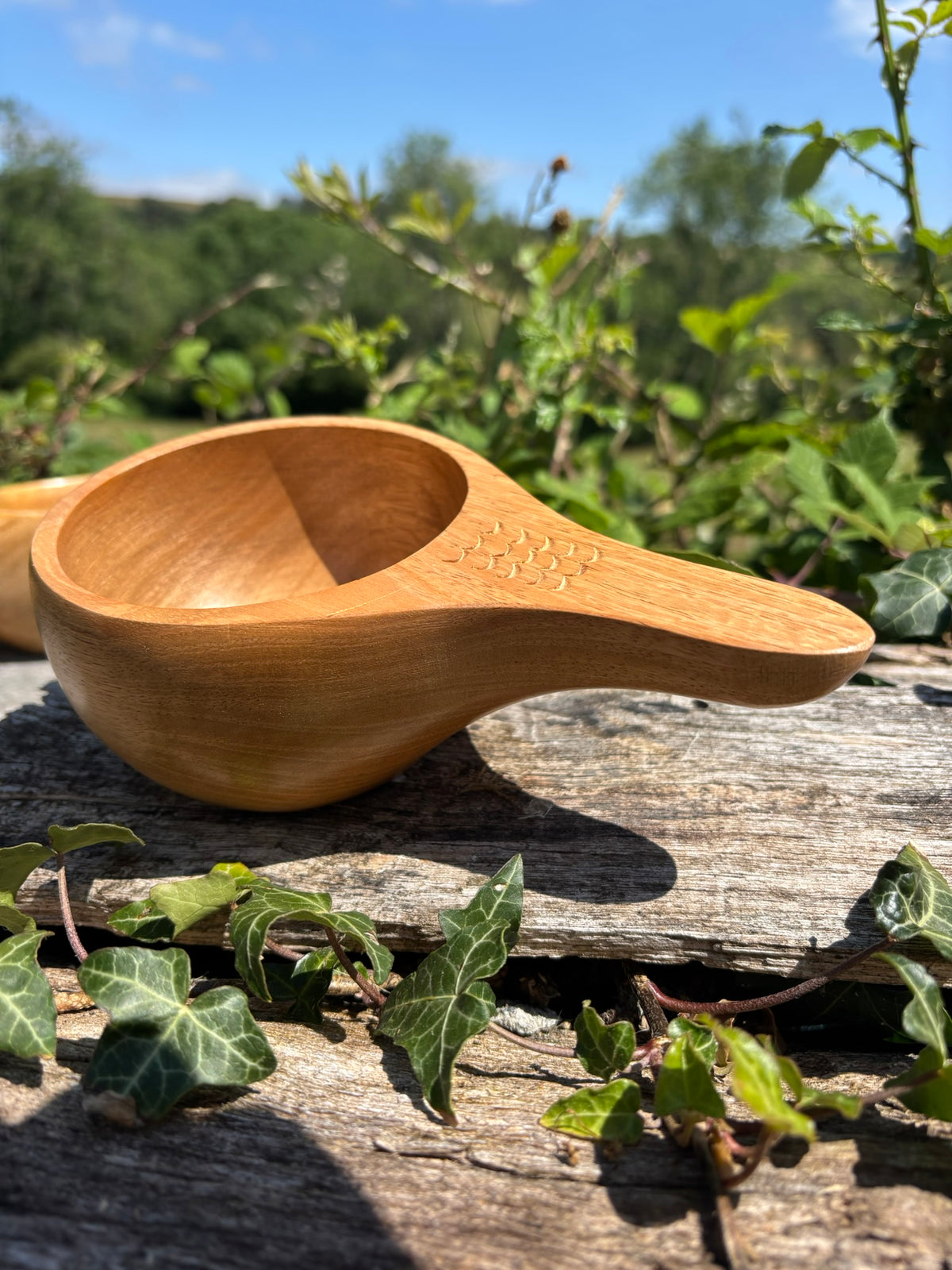 A small handcrafted wooden Kuksa Cup made from light natural wood, resting on a weathered log outdoors with green ivy and a blurred garden background.