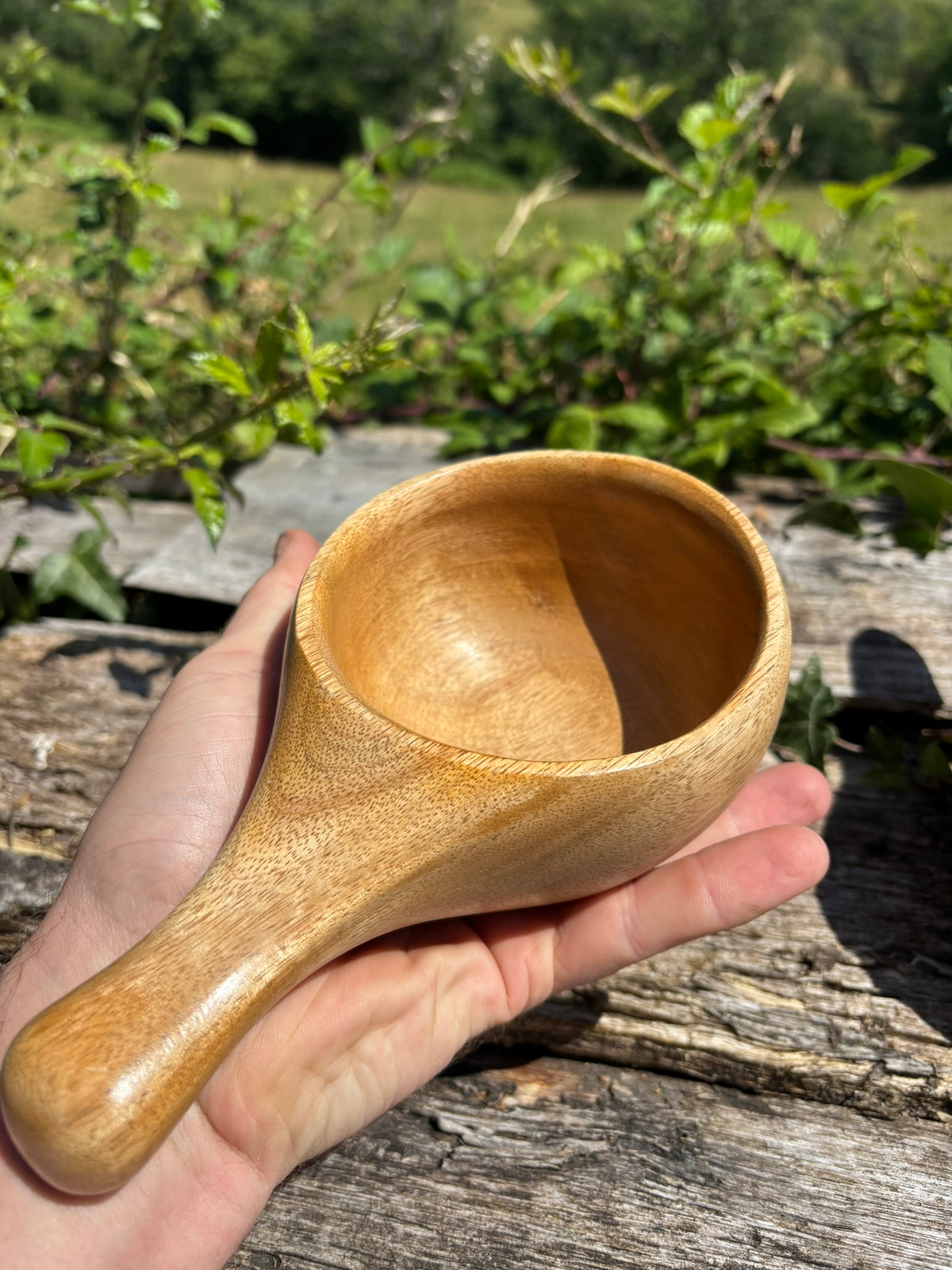 A Viking Cup or Kuksa, with a rustic weathered wood surface and green foliage blurred in the background.