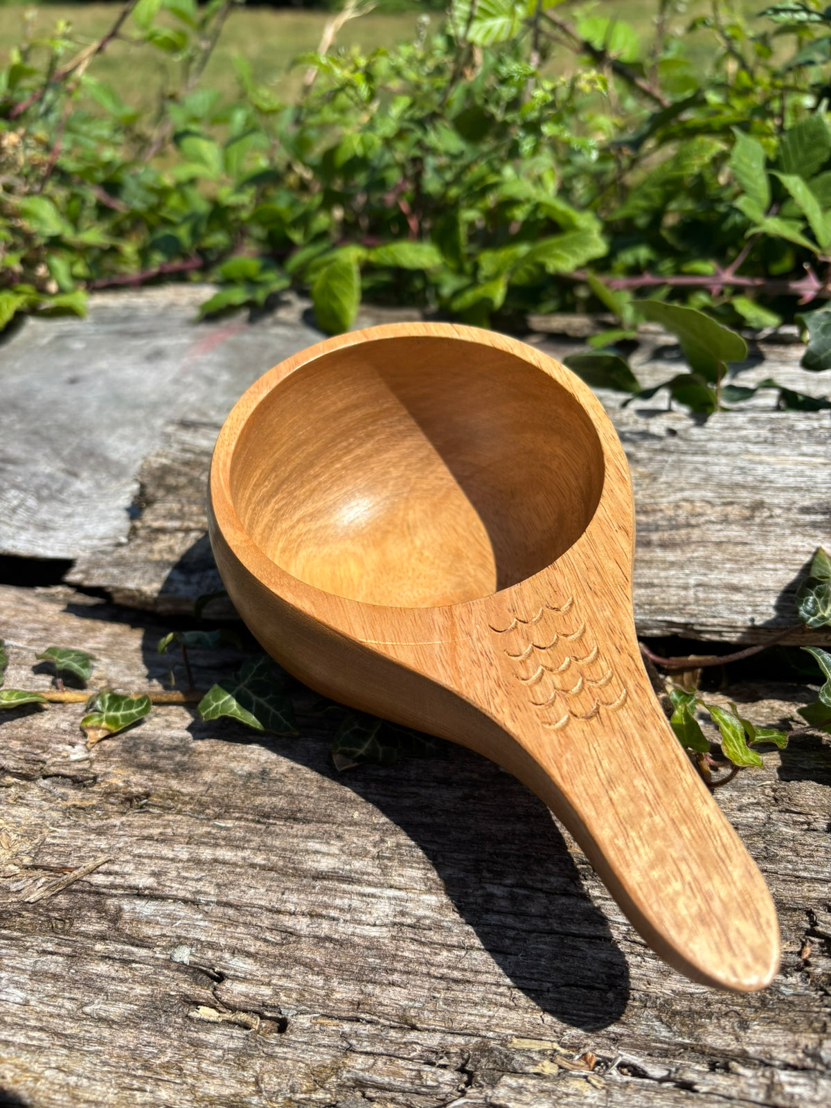 A small handcrafted Kuksa with a fish-scale pattern carved on the handle, resting on a weathered wooden surface outdoors with green foliage in the background.
