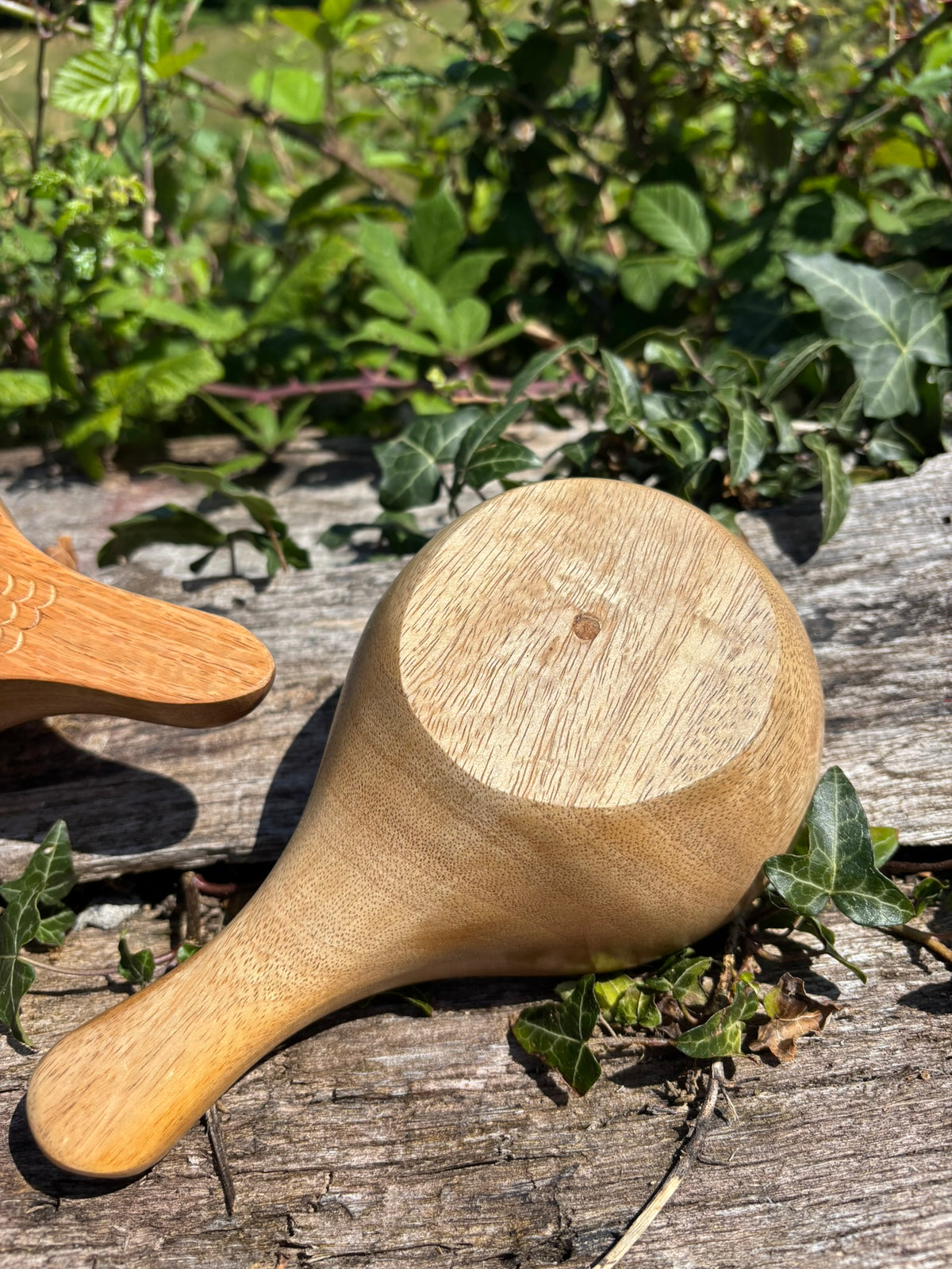 A hand turned Kuksa showing the turning spot, with a rounded bowl and short handle, photographed outdoors on a weathered wooden surface with green ivy and foliage in the background.