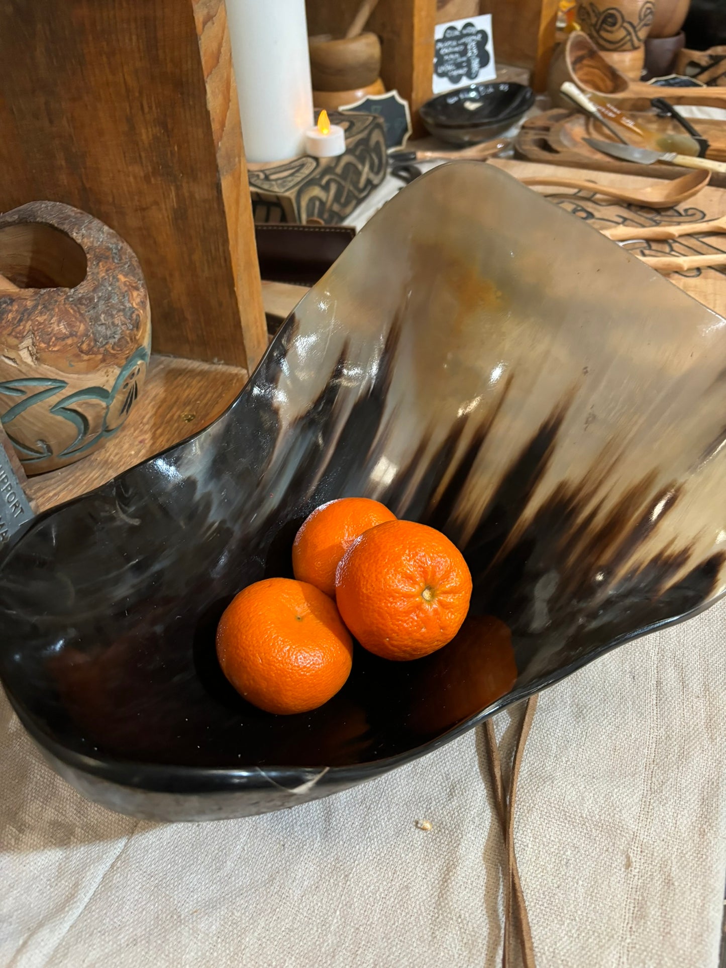 A dark, organic-shaped horn bowl with a glossy black and brown marbled finish, containing three oranges, set on a beige linen tablecloth in a rustic, handcrafted shop environment.
