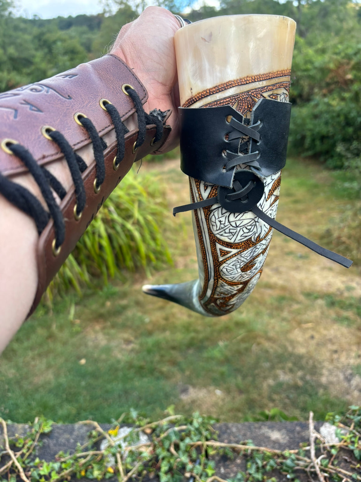 A hand holding a carved horn cup with a brown leather sleeve and black laces, set against an outdoor grassy background.