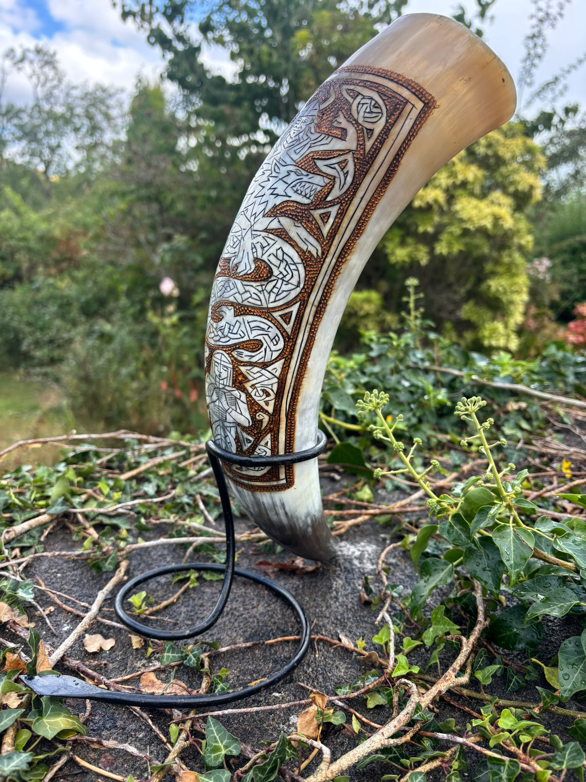 A carved horn or tusk-shaped decorative object with intricate Celtic-style knotwork and geometric engravings, displayed outdoors on a black metal stand among green plants and branches.