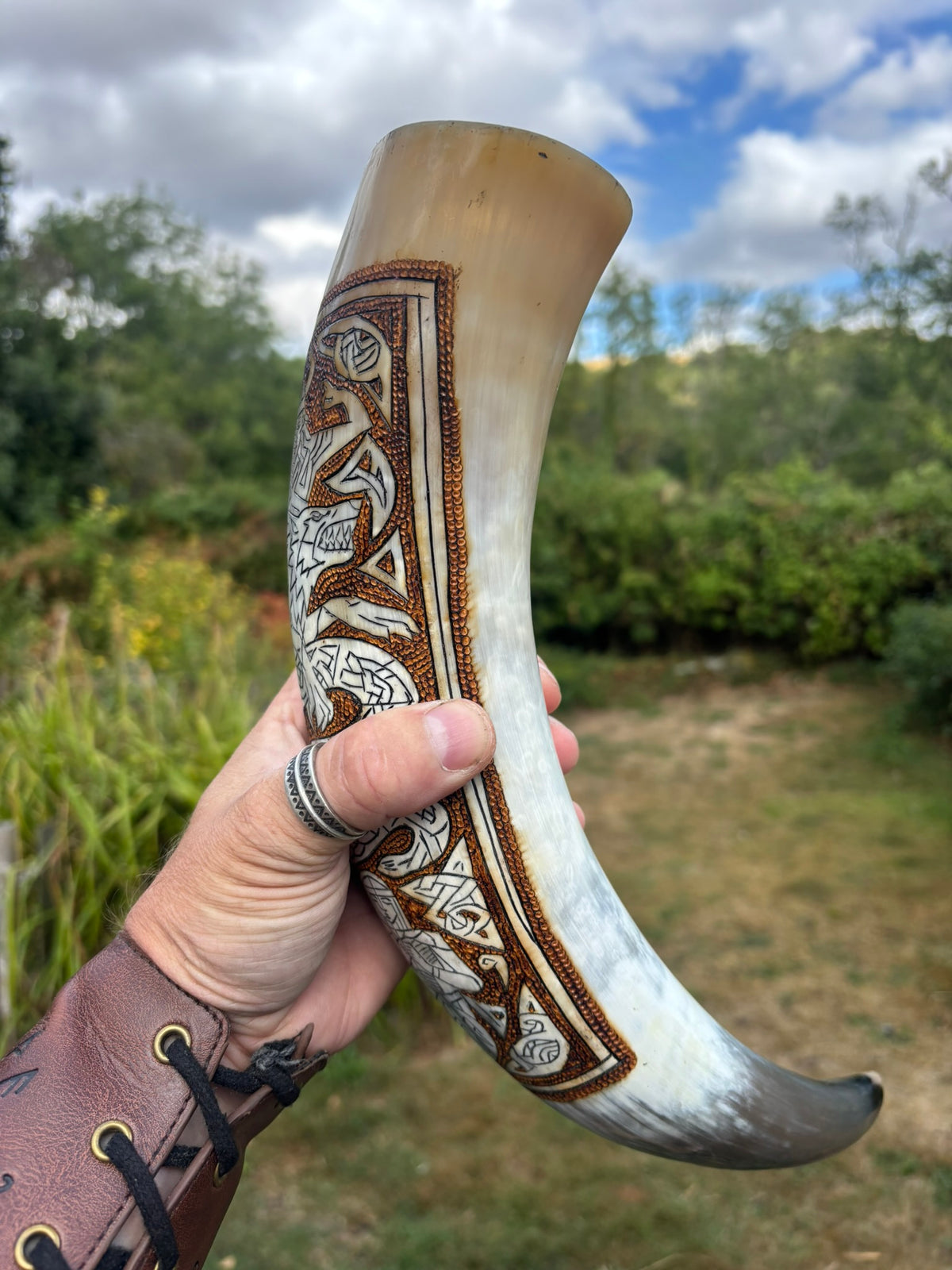 A hand holds a carved horn cup outdoors, featuring intricate brown and white Celtic knotwork and geometric engravings on the side. The horn has a natural beige top and a dark gray curved bottom, with a blurred green and brown outdoor background.