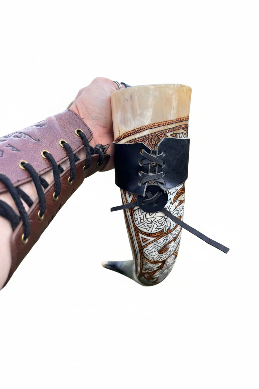 A hand holding a carved horn or bone drinking cup decorated with brown and silver patterns, wrapped in a black lace-up band, with a brown leather arm guard visible on the wrist against an outdoor grassy background.