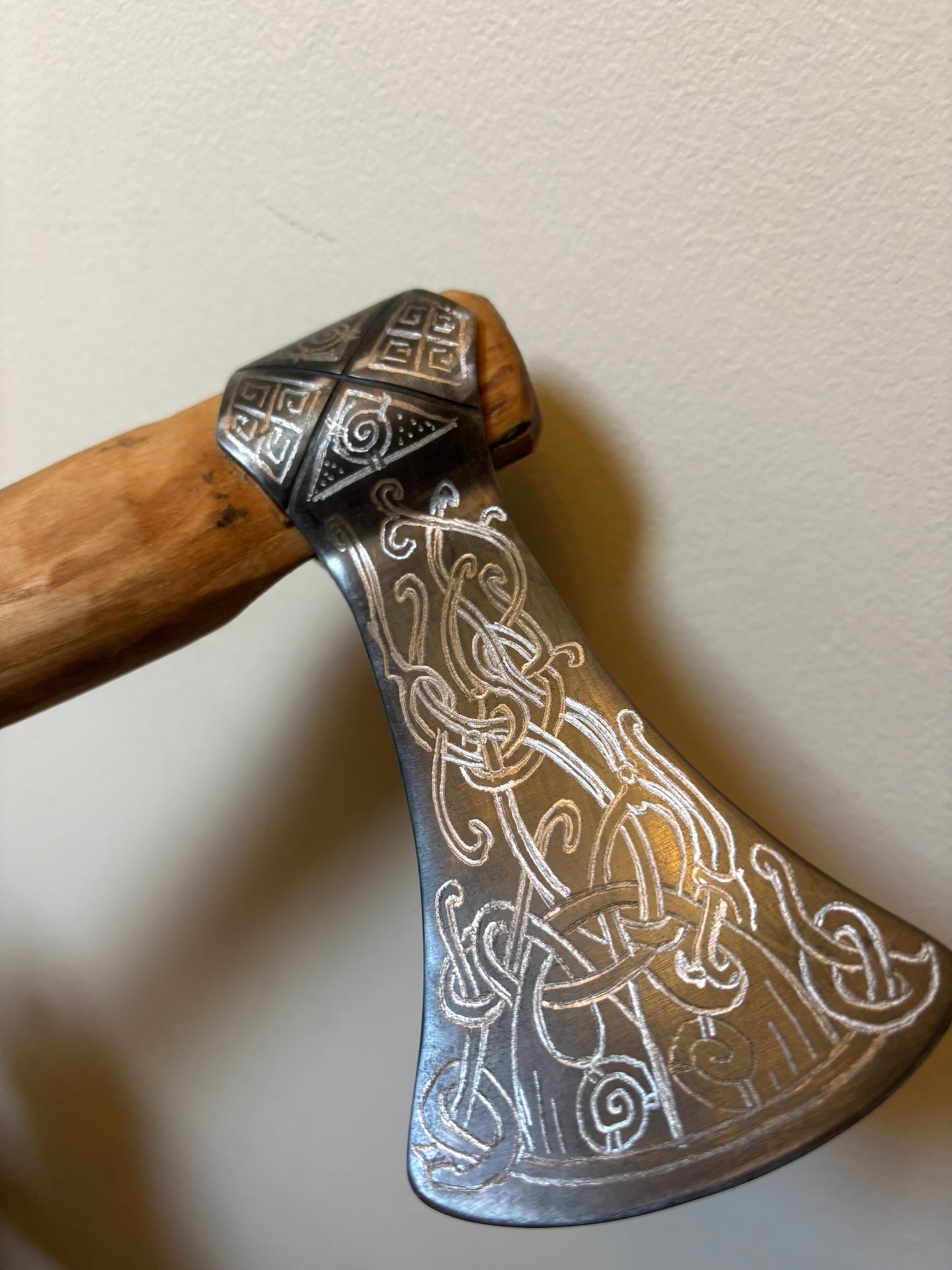 A close-up of a decorative axe or hatchet with an engraved silver-toned metal head featuring intricate Celtic knotwork patterns, attached to a wooden handle, set against a plain beige wall.