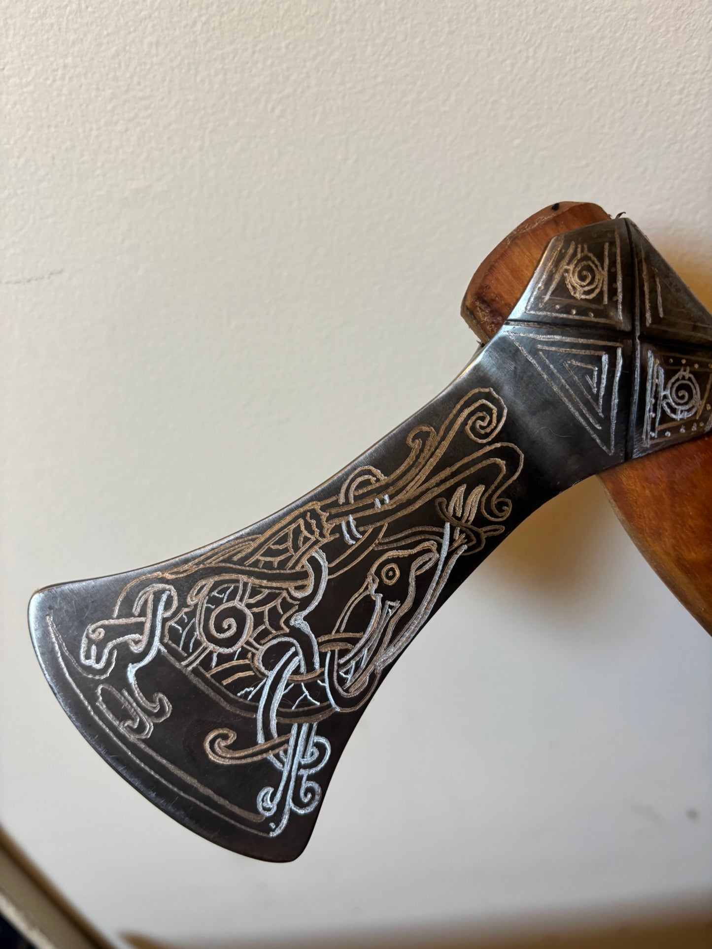 A close-up of a decorative Mammen Axe head with engraved Nordic-style knotwork patterns in silver and gold tones on a dark metal surface, with part of a wooden handle visible against a plain off-white wall.
