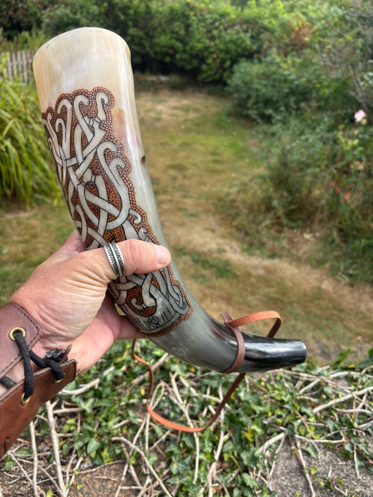 A Viking Holds a Mammen Carved Blowing Horn, brown and white Celtic knotwork patterns, featuring a dark metal mouthpiece and leather straps, set against an outdoor garden background.
